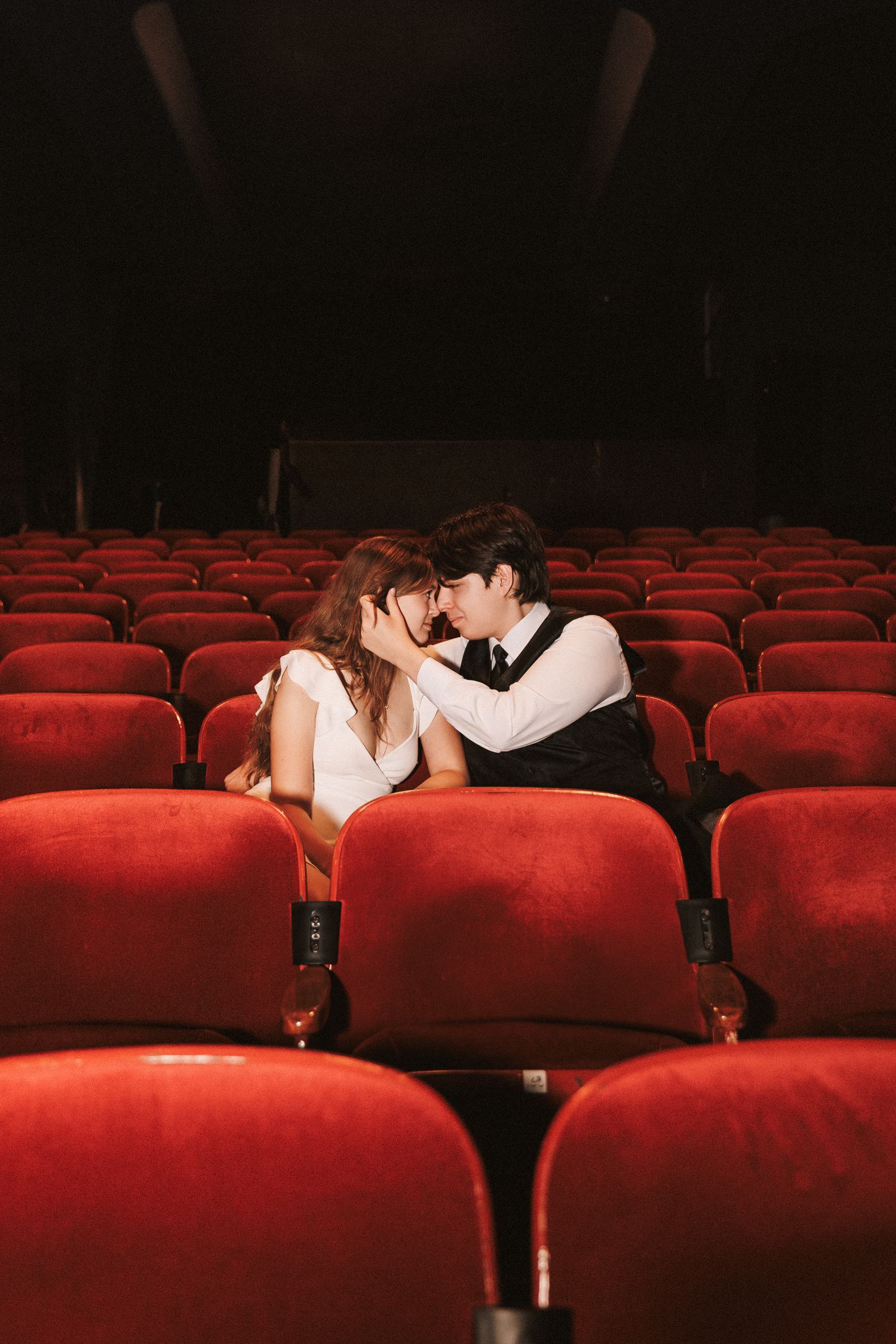 A romantic couple shares an intimate kiss in the empty red seats of a darkened movie theater or auditorium.
