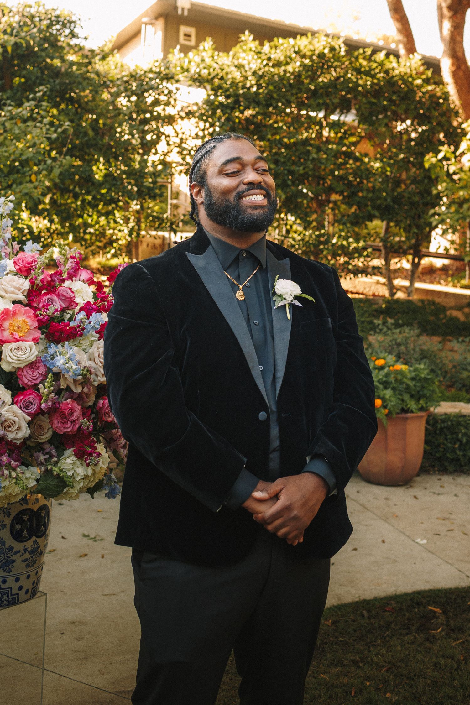 Joyful groom in black velvet blazer laughing beside colorful floral arrangement at outdoor ceremony.