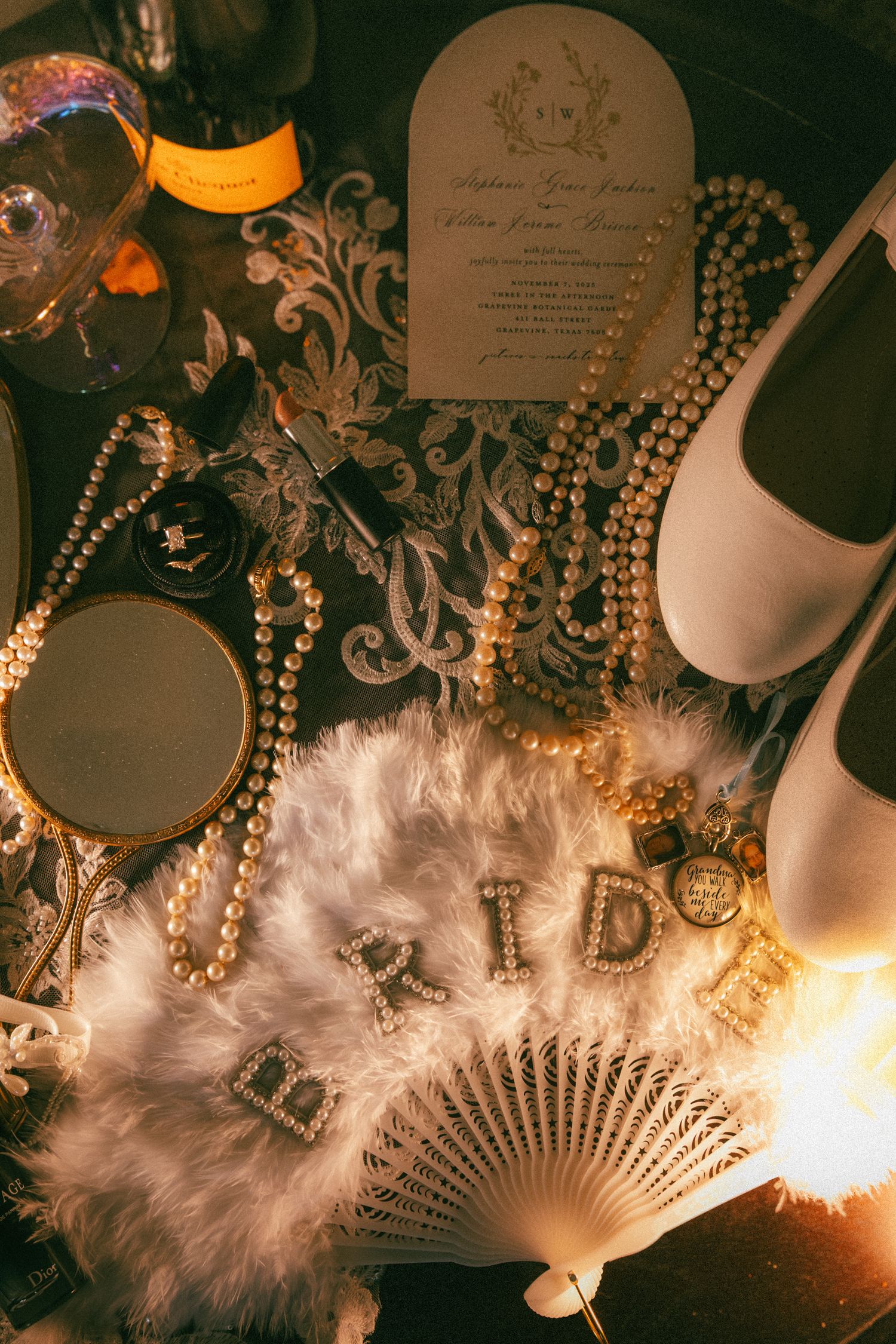 Elegant bridal flat lay with pearl necklaces, feather fan, white heels, and wedding invitation on dark floral background.