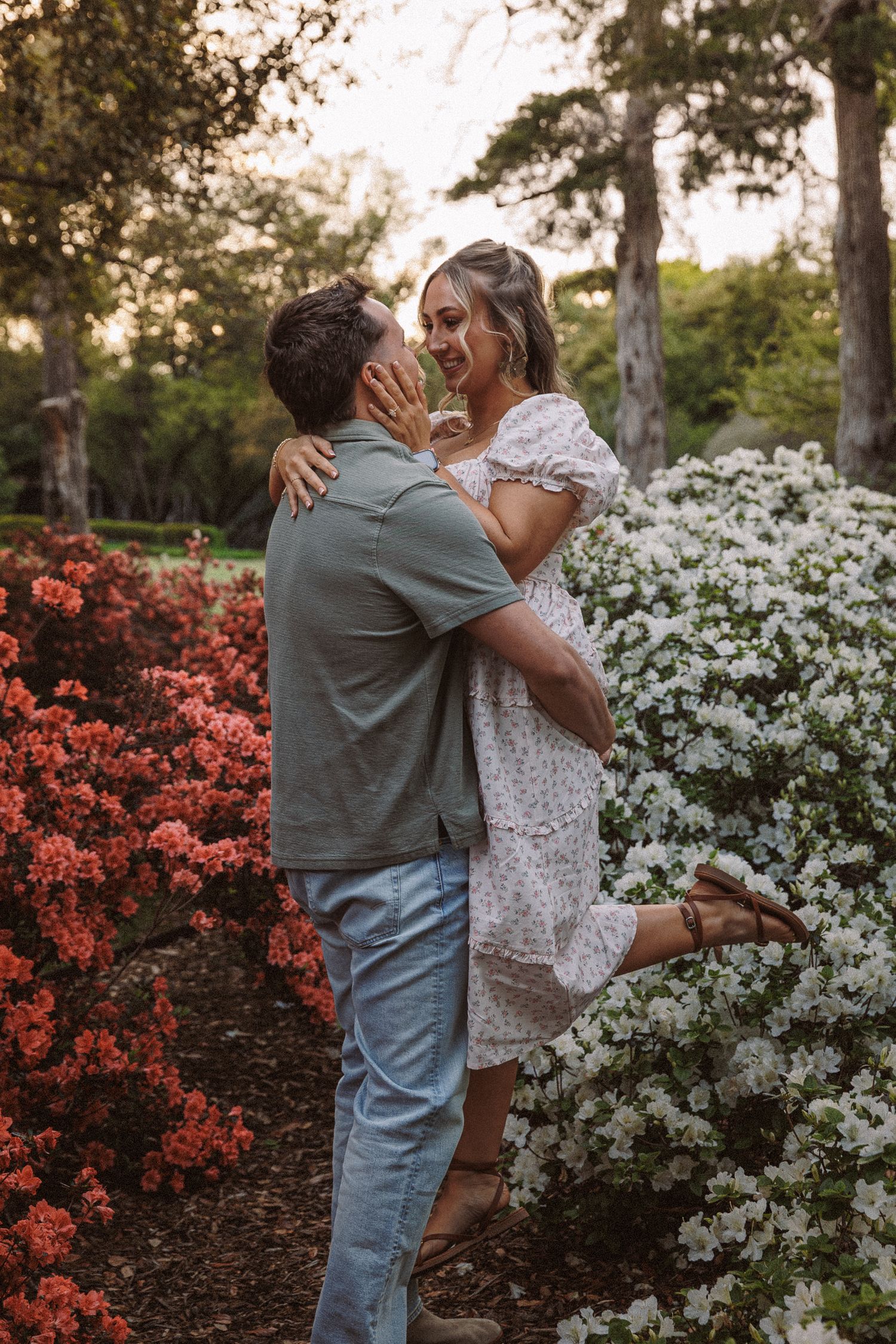 Couple embracing among red and white flowering bushes in a lush garden at golden hour.