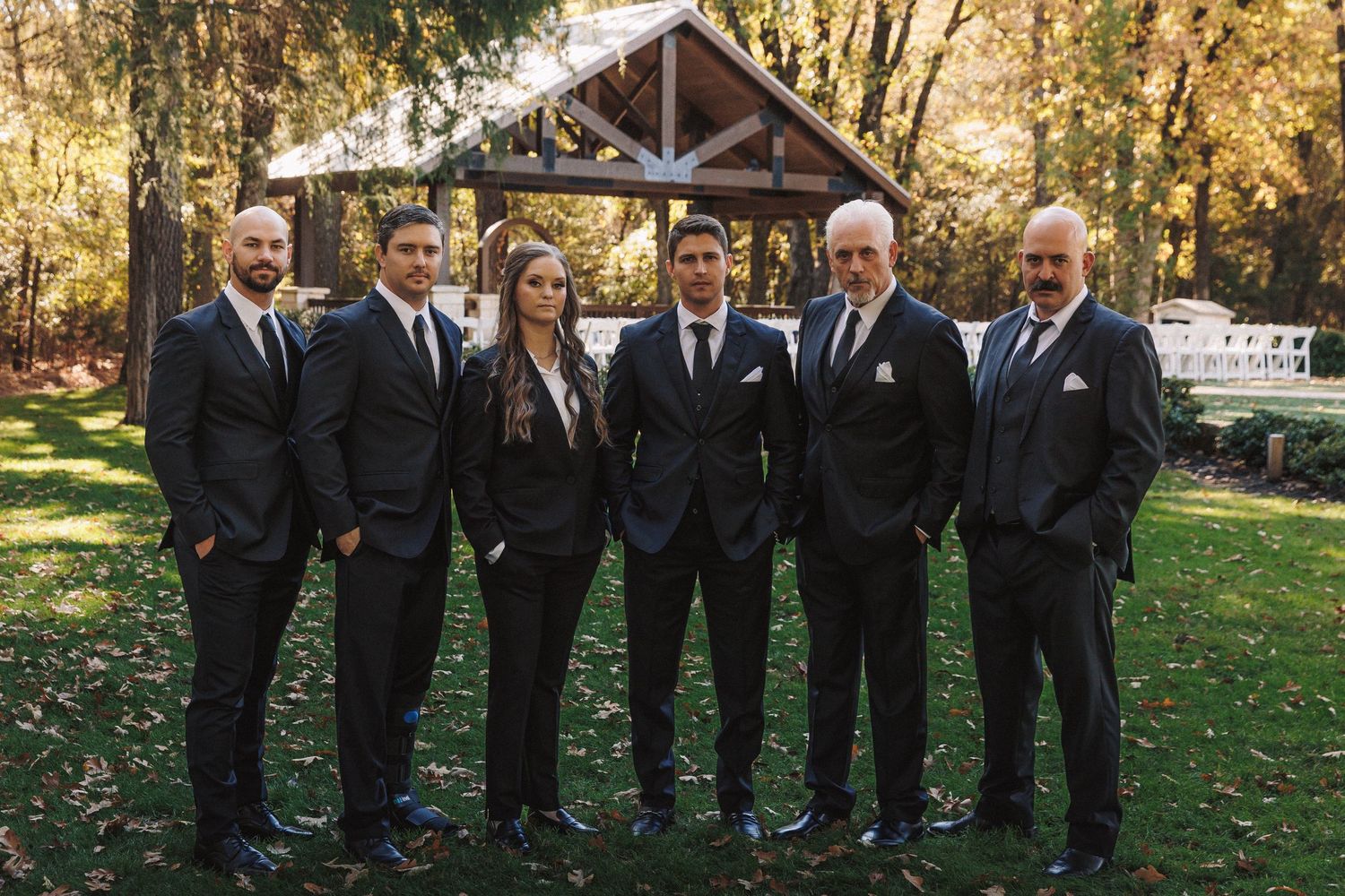 Six groomsmen in matching black suits posing together outdoors near a wooden wedding ceremony pavilion.