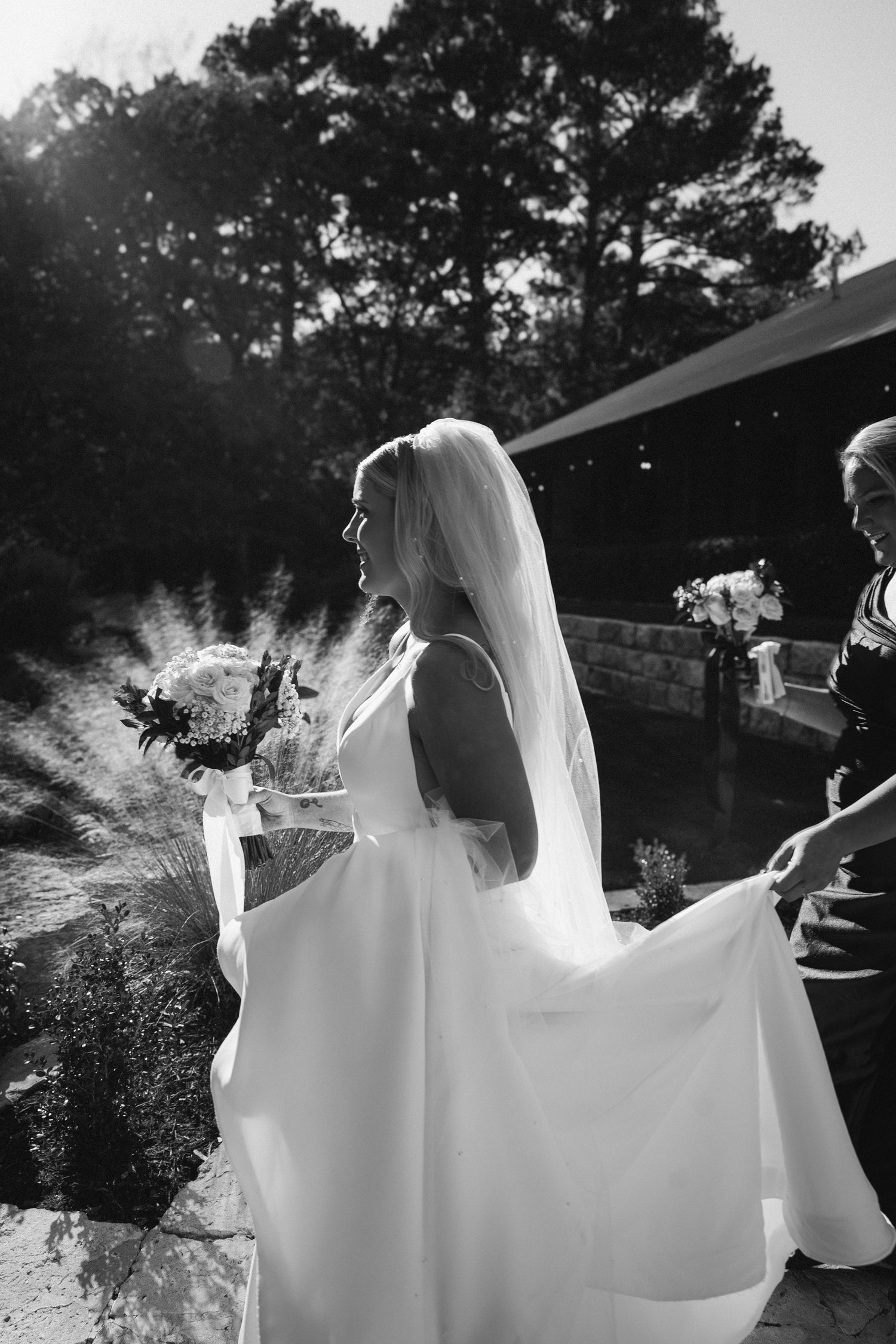 Black and white photo of a bride in a flowing white gown and veil holding a bouquet outdoors.