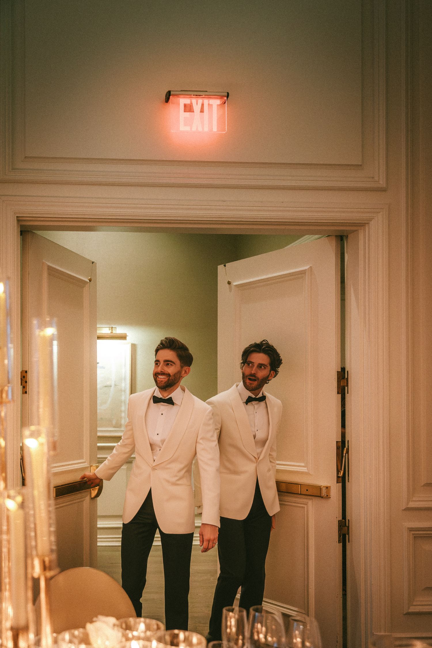 Two grooms in white tuxedos and bow ties making grand entrance through reception doors.