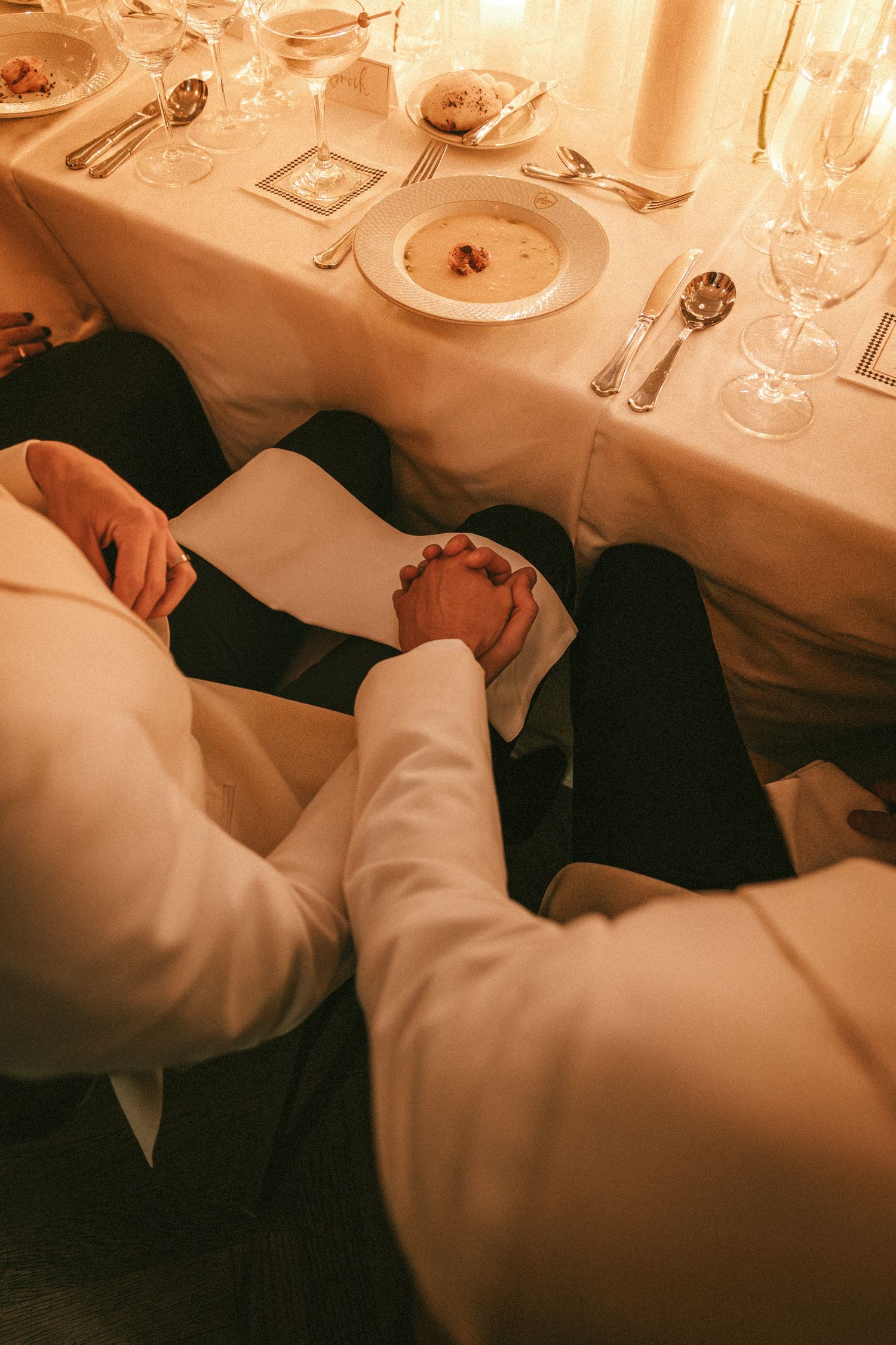 Two grooms in white suits hold hands under elegant wedding reception dinner table.