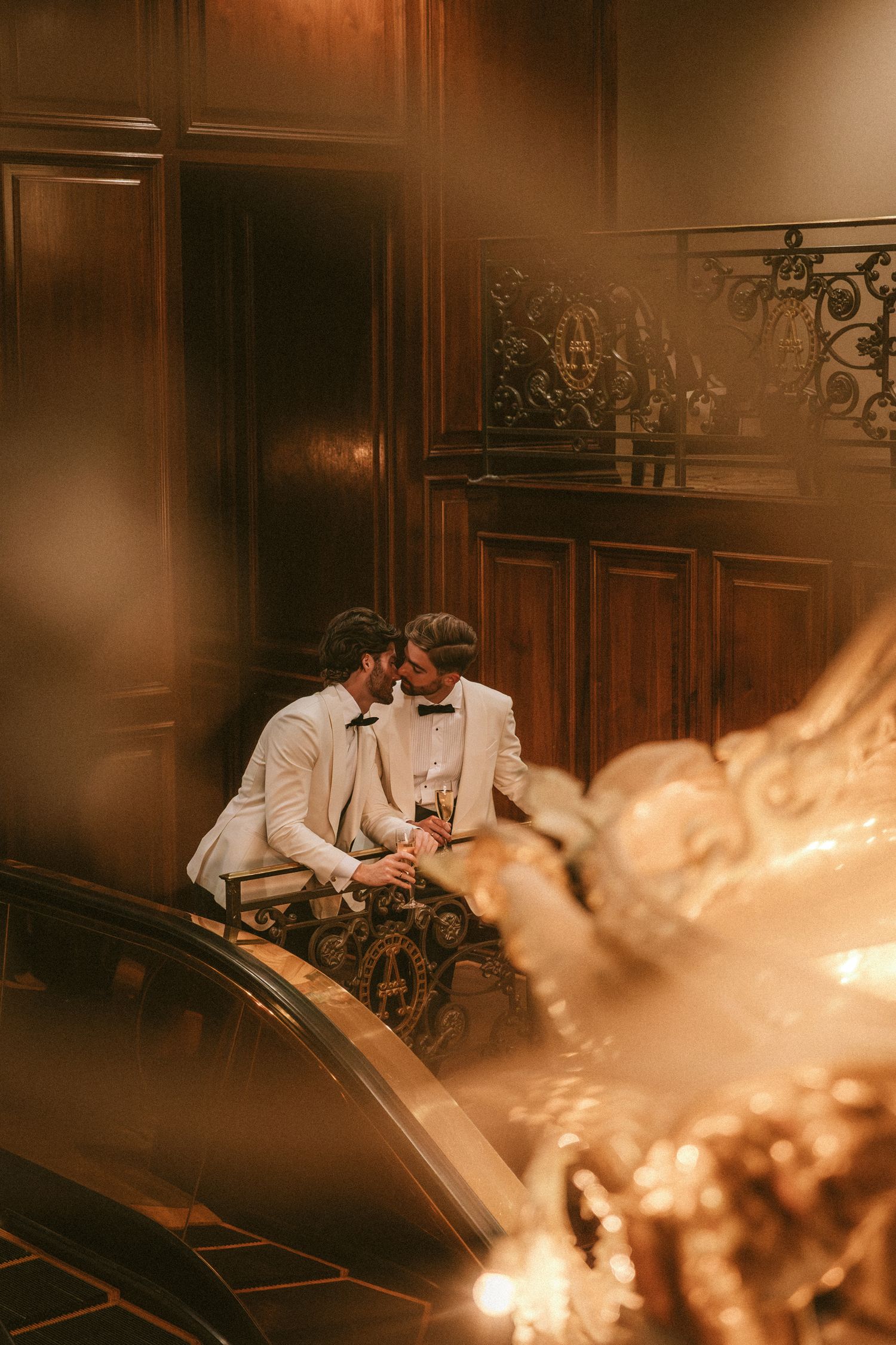 Two grooms share an intimate moment on a grand staircase in a warmly lit venue.