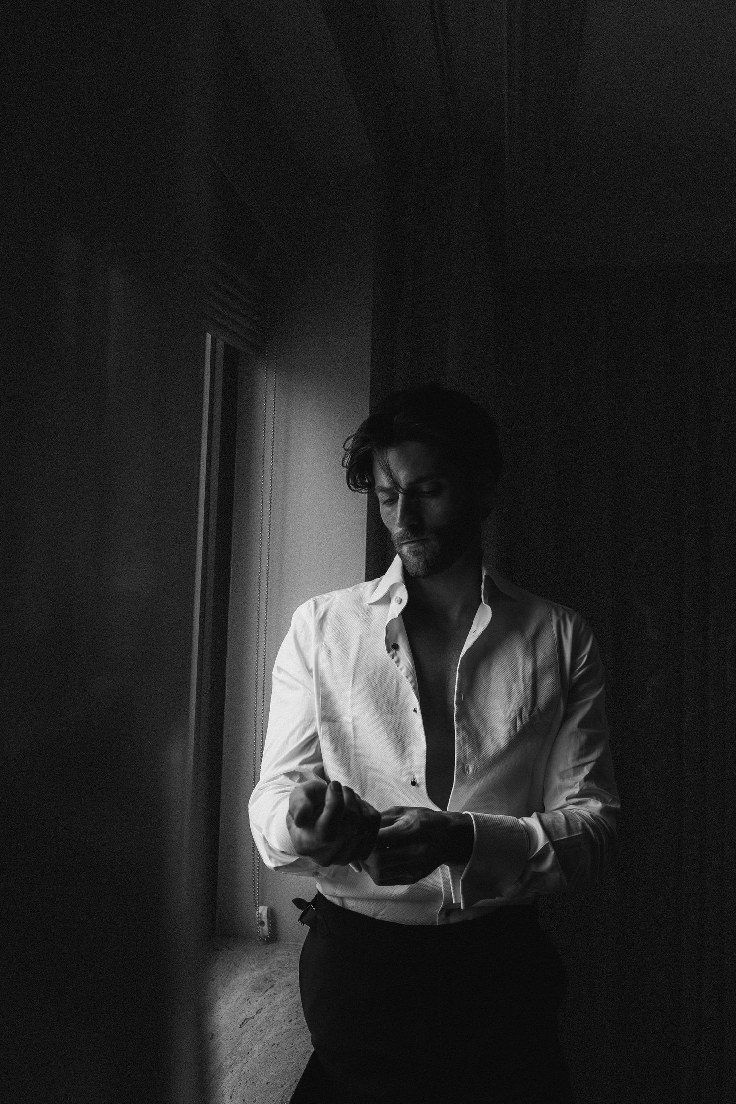 Black and white portrait of a groom in a white dress shirt adjusting his cufflinks in dramatic window light.