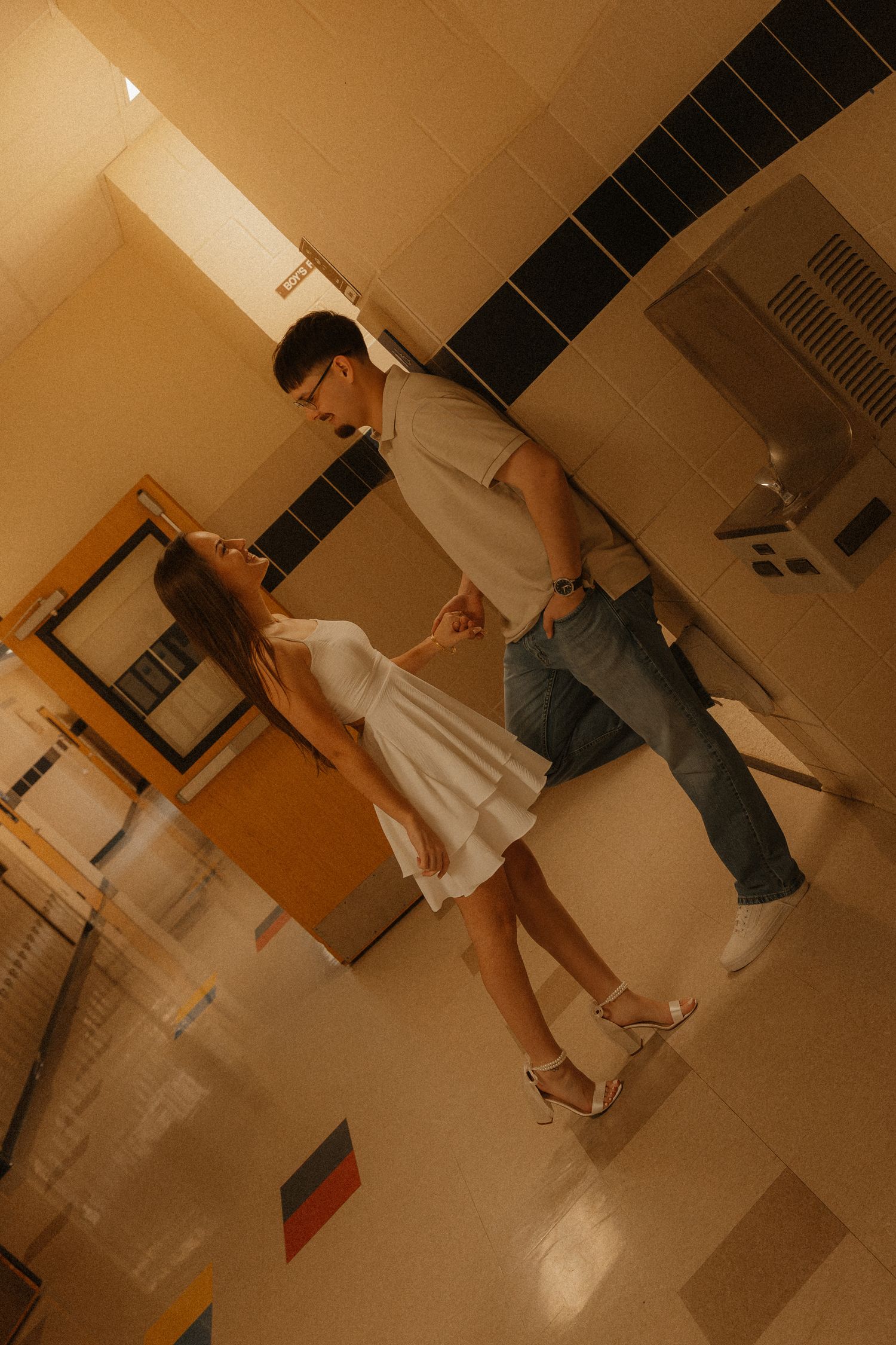 Couple dancing together in a warmly lit school hallway, woman in white dress and heels.