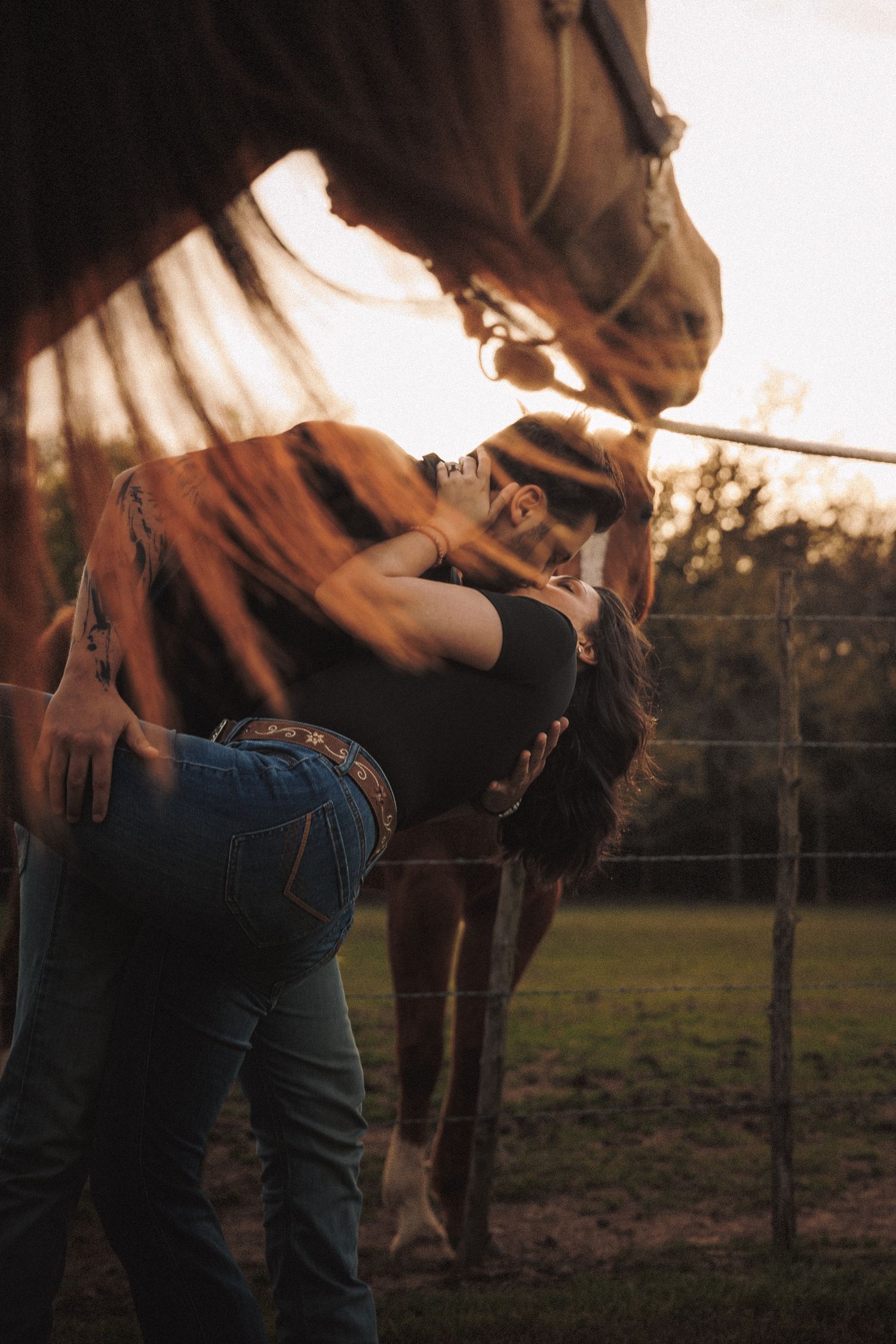 Romantic couple kissing beside a brown horse along a fence at warm sunset.