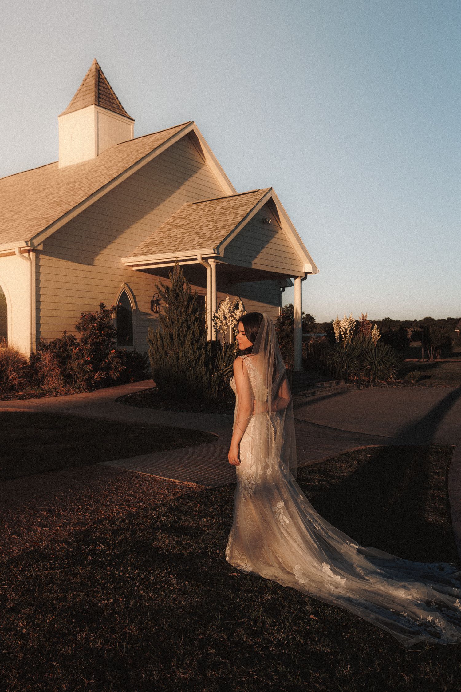 Bride in lace wedding gown with long veil standing outside a white chapel at golden hour.