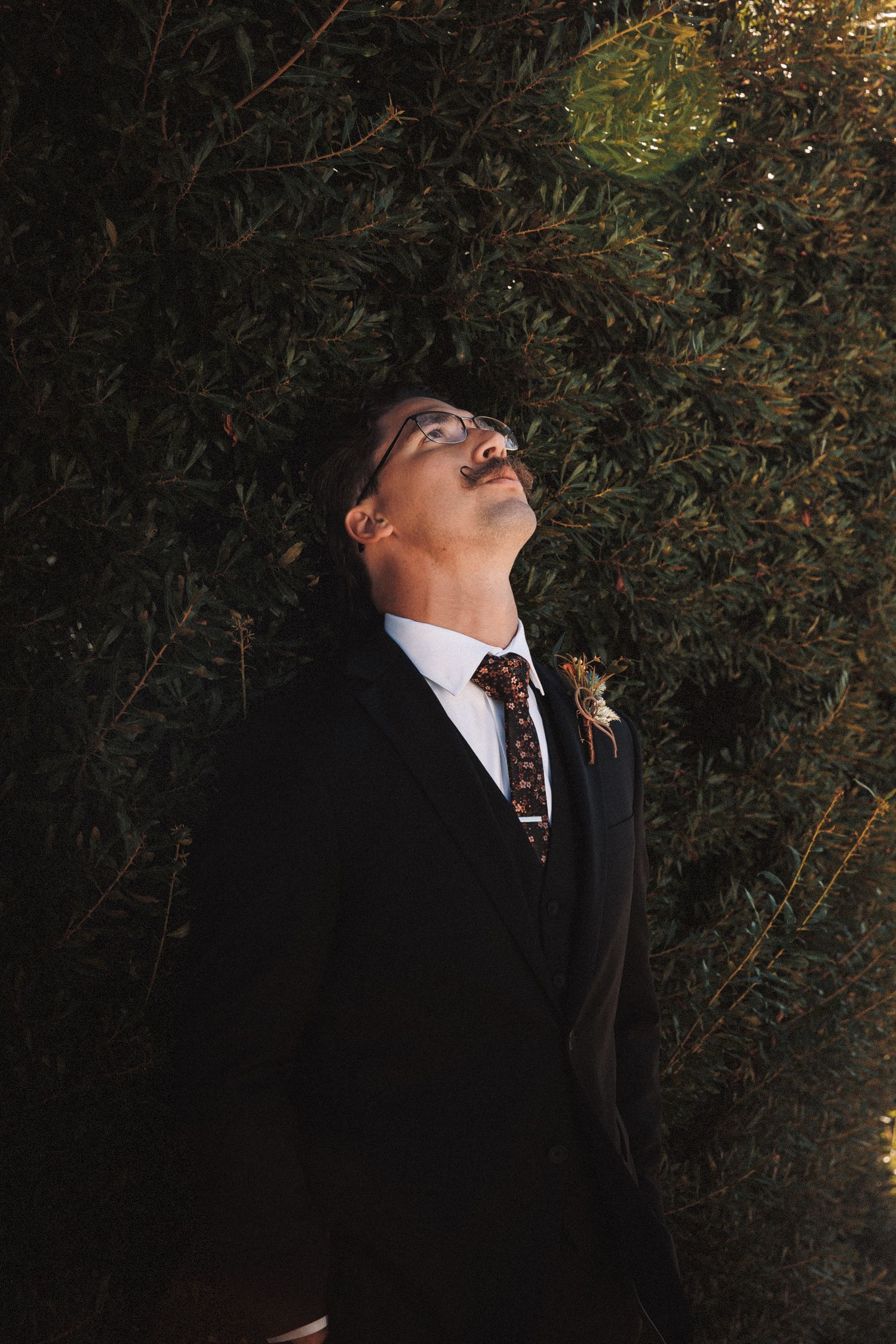 Groom in black suit with floral tie and boutonniere tilts head back near lush green bushes at night.