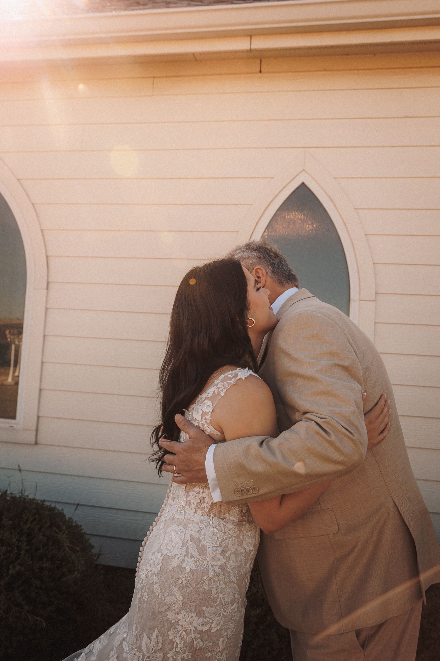 Father of the bride shares emotional first look hug outside sunlit white chapel.