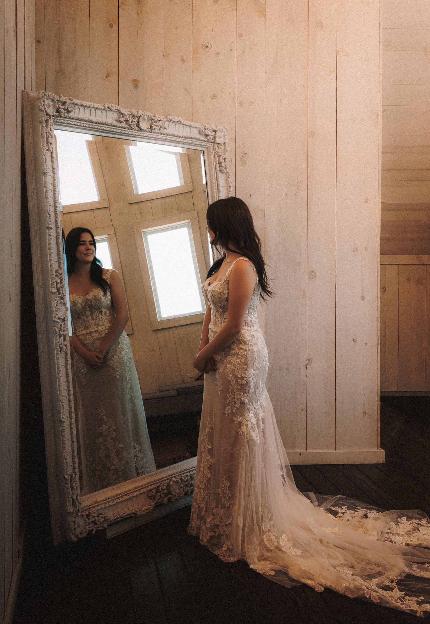 Bride in lace wedding gown admiring her reflection in an ornate white mirror in a rustic wood-paneled room.