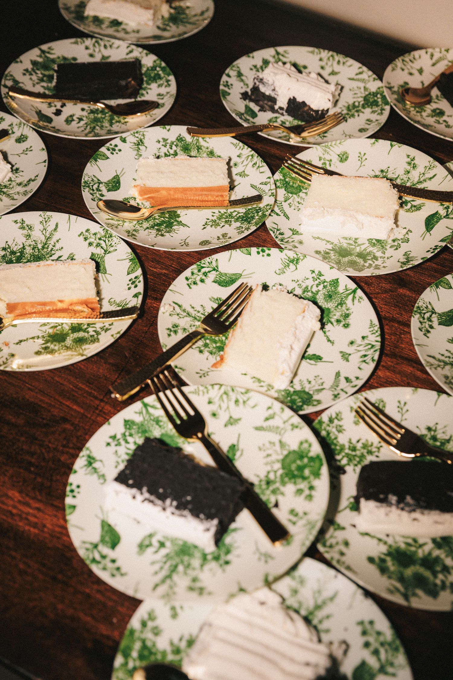 Wedding cake slices served on green floral plates with gold forks on wooden table.
