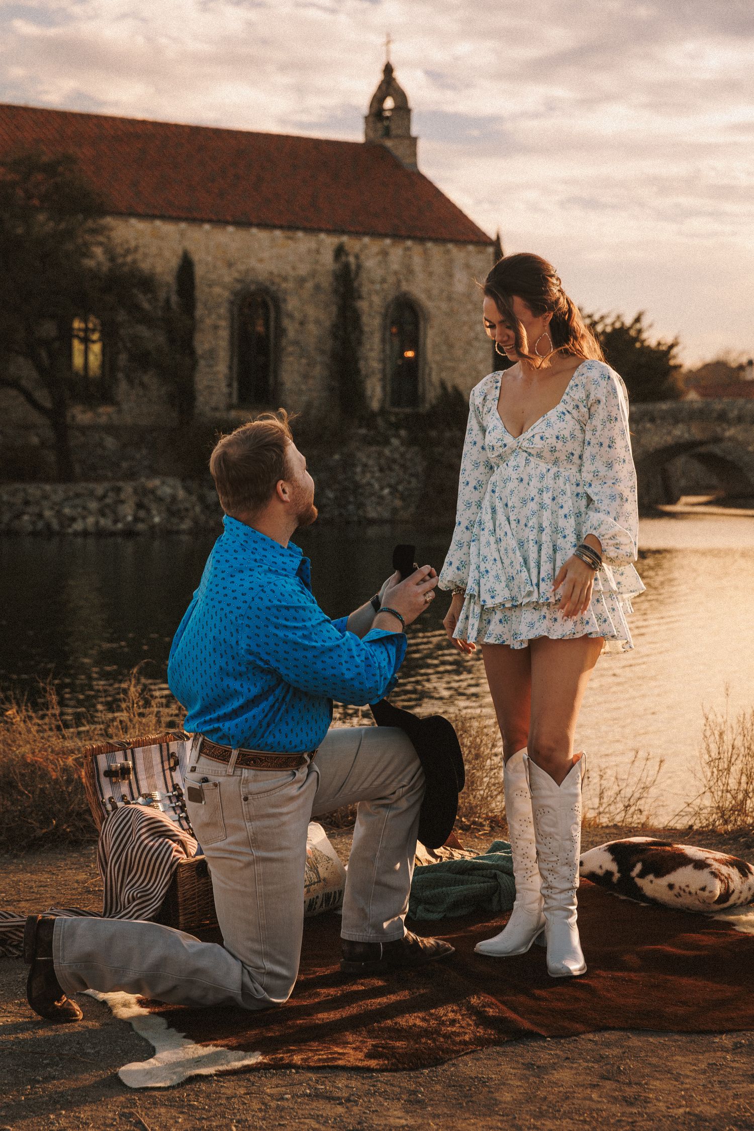A romantic marriage proposal scene at sunset by a waterfront with a couple in casual white and blue outfits.