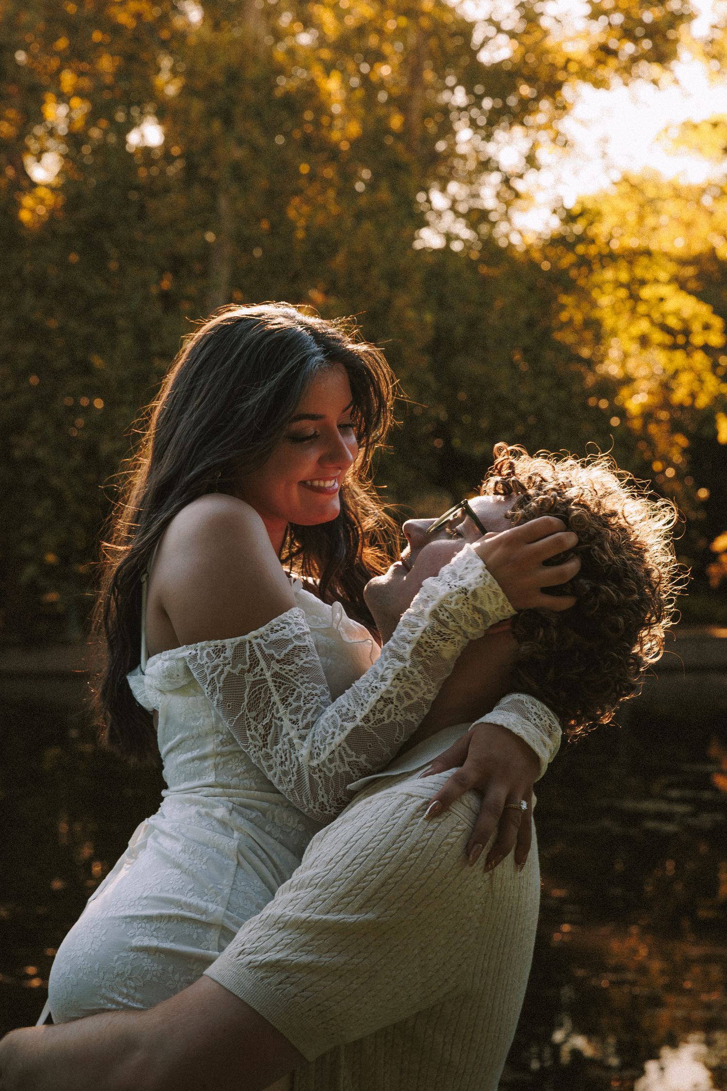 Romantic couple embraces in a white lace dress during golden hour sunset in a park setting.