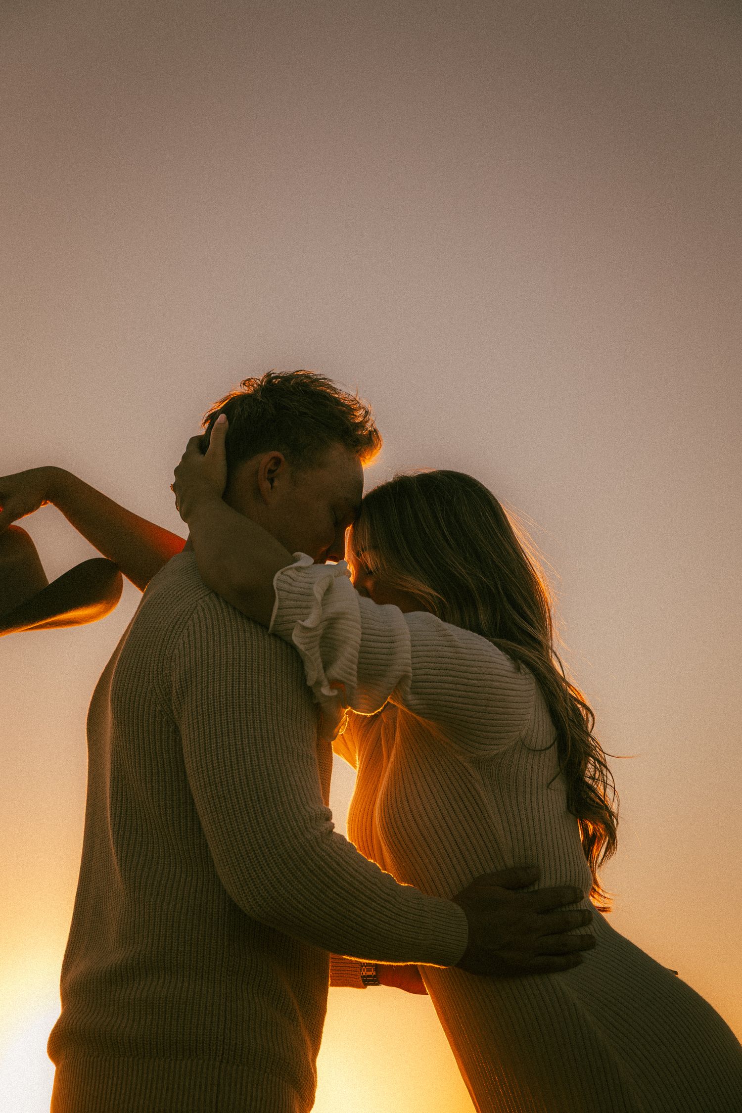 Silhouetted couple in sweaters embracing during golden hour with warm backlighting.