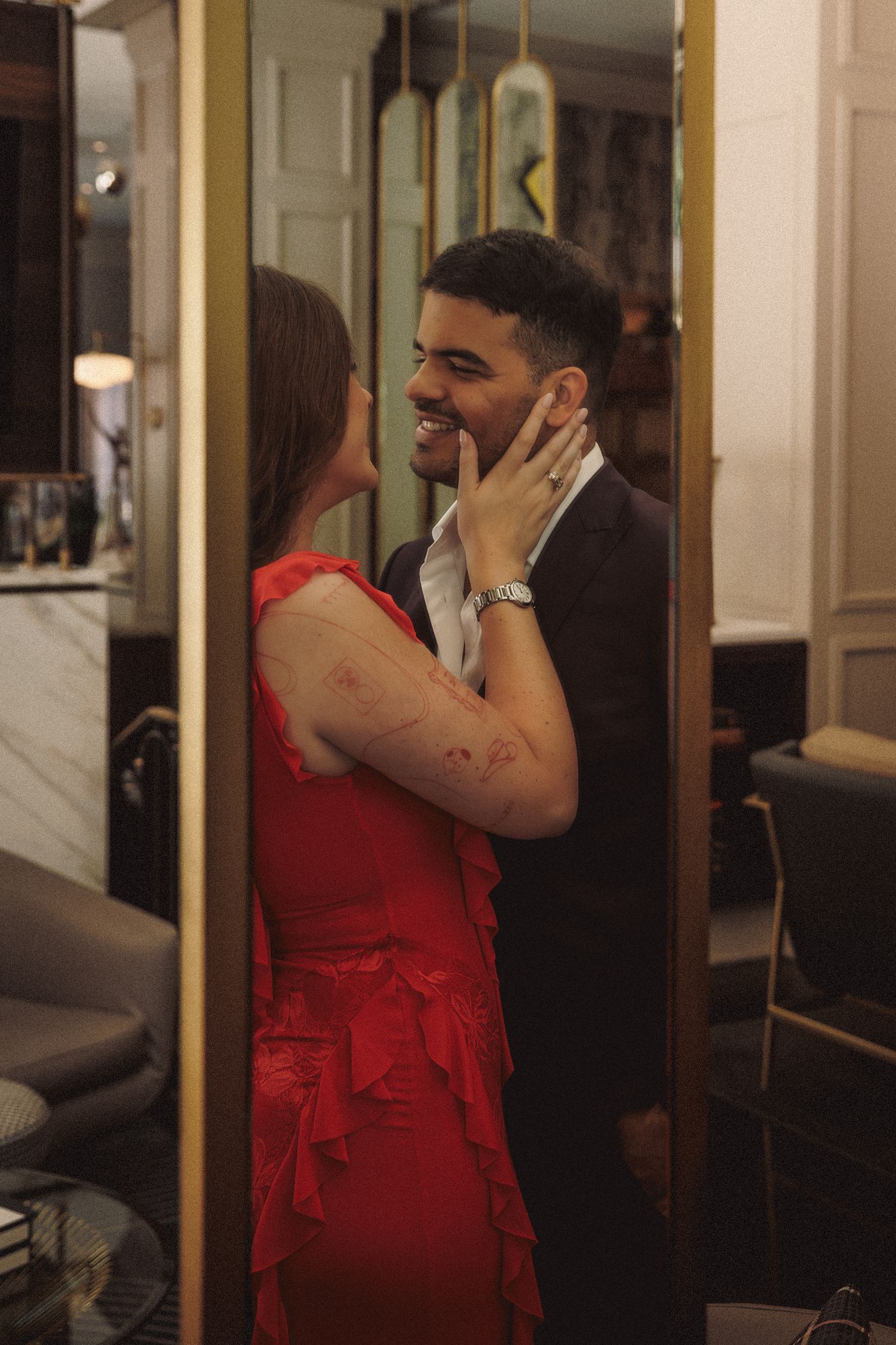 Couple sharing a romantic moment, woman in red dress tenderly holding mans face while he smiles in elegant interior.