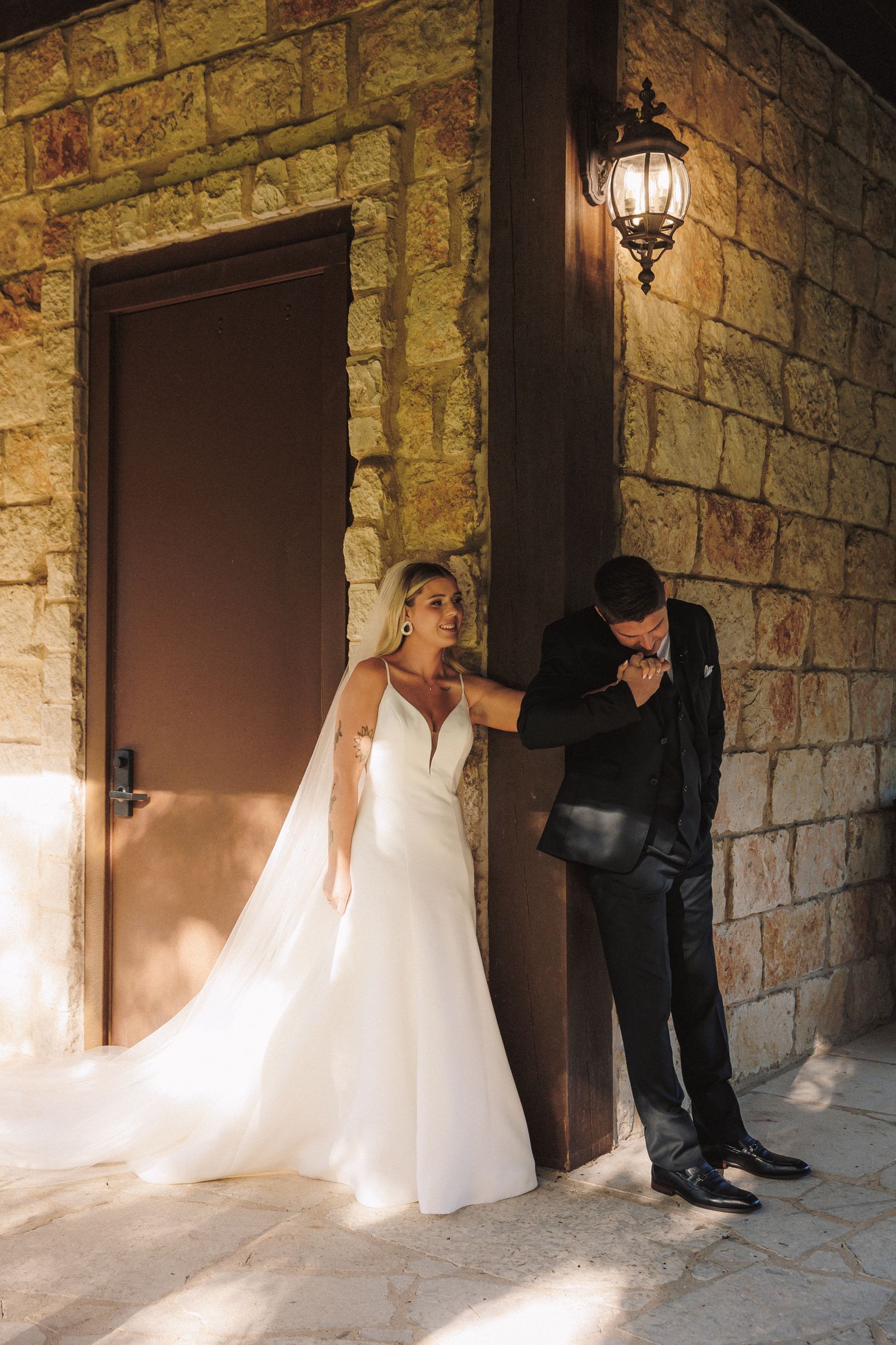 Groom kissing bride's hand against stone wall with vintage lantern at elegant wedding venue.