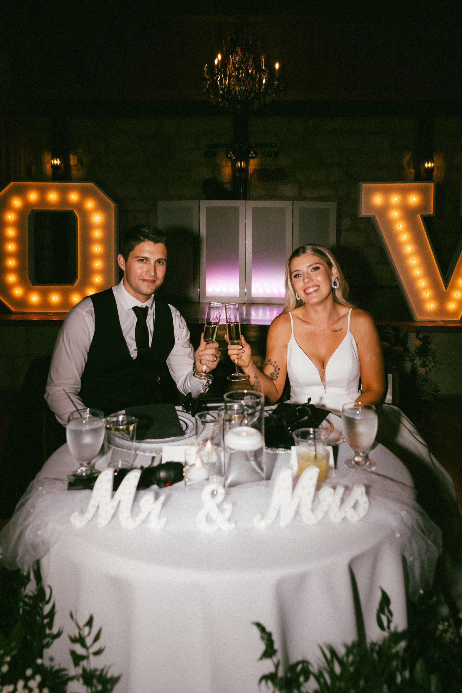 Newlyweds toast with champagne at sweetheart table decorated with Mr and Mrs marquee sign.