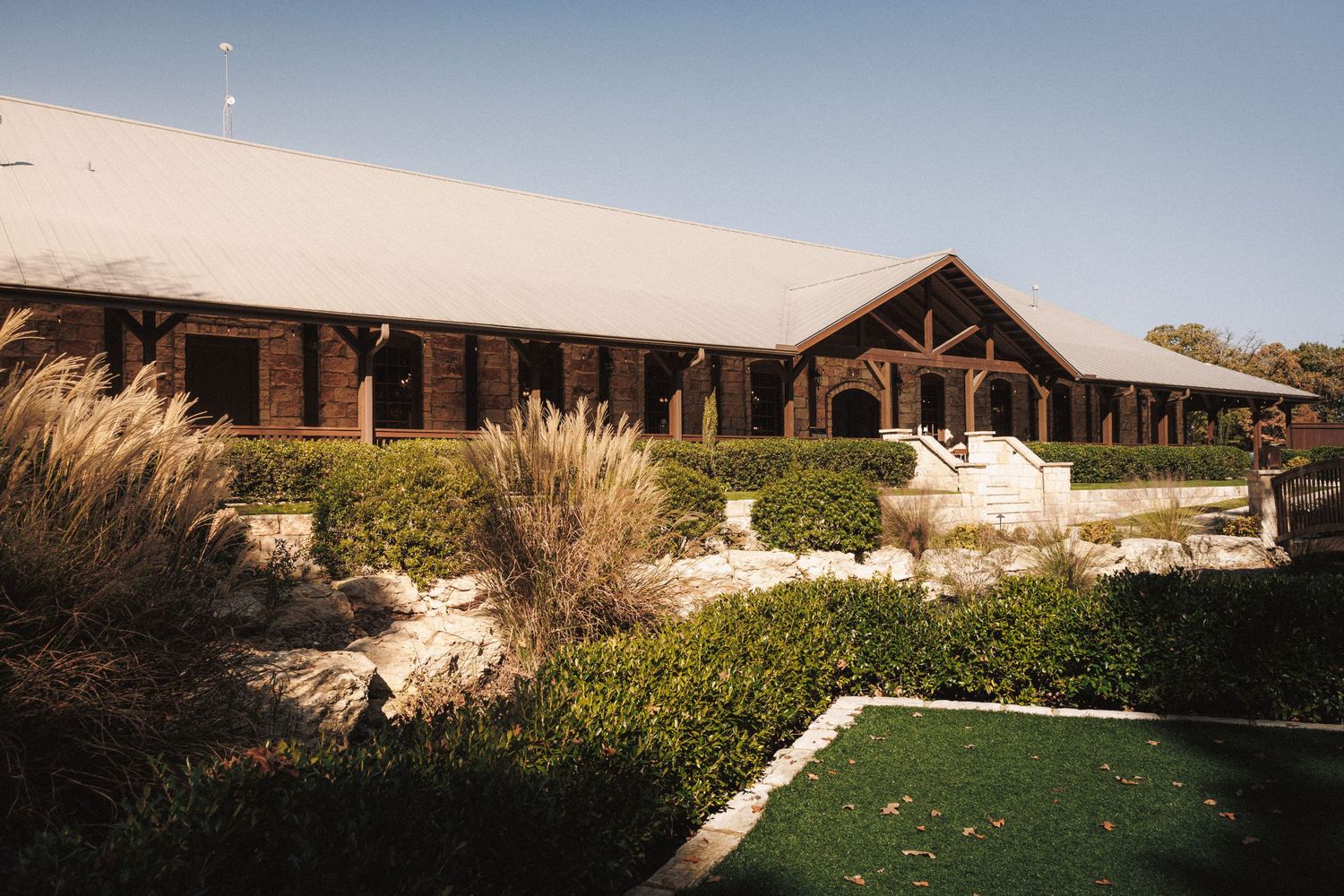 Rustic wooden wedding venue exterior with manicured hedges and ornamental grasses under a clear blue sky.