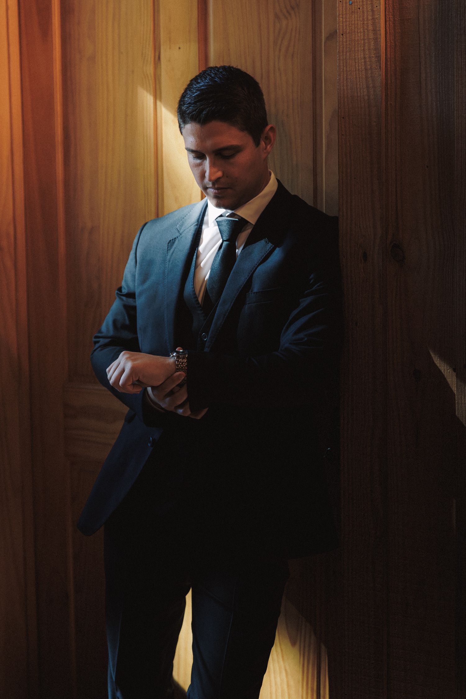 Handsome groom in dark suit checking his watch while leaning against wooden paneled wall in warm light.