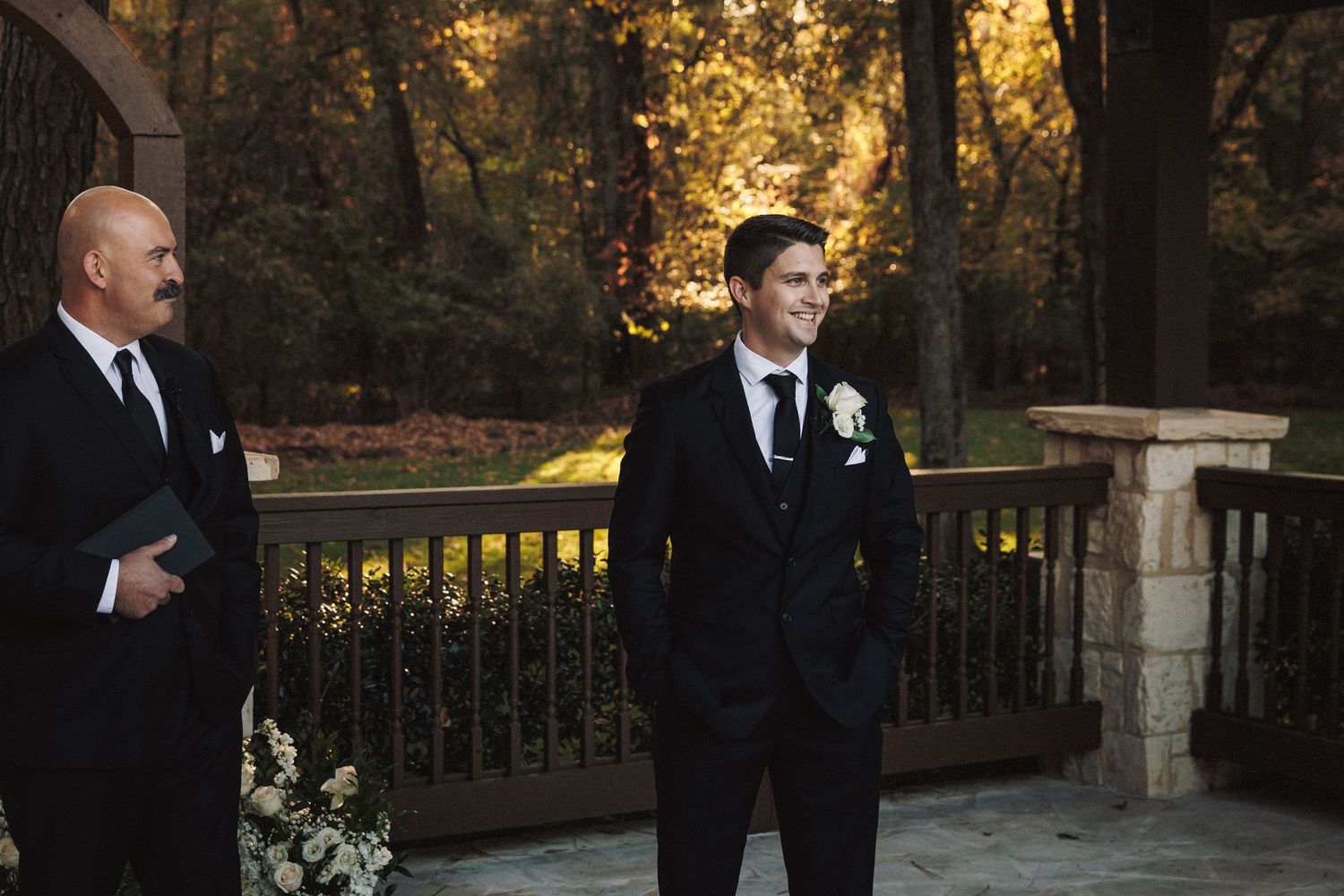 Groom in black suit with white boutonniere stands at outdoor wedding altar with officiant.