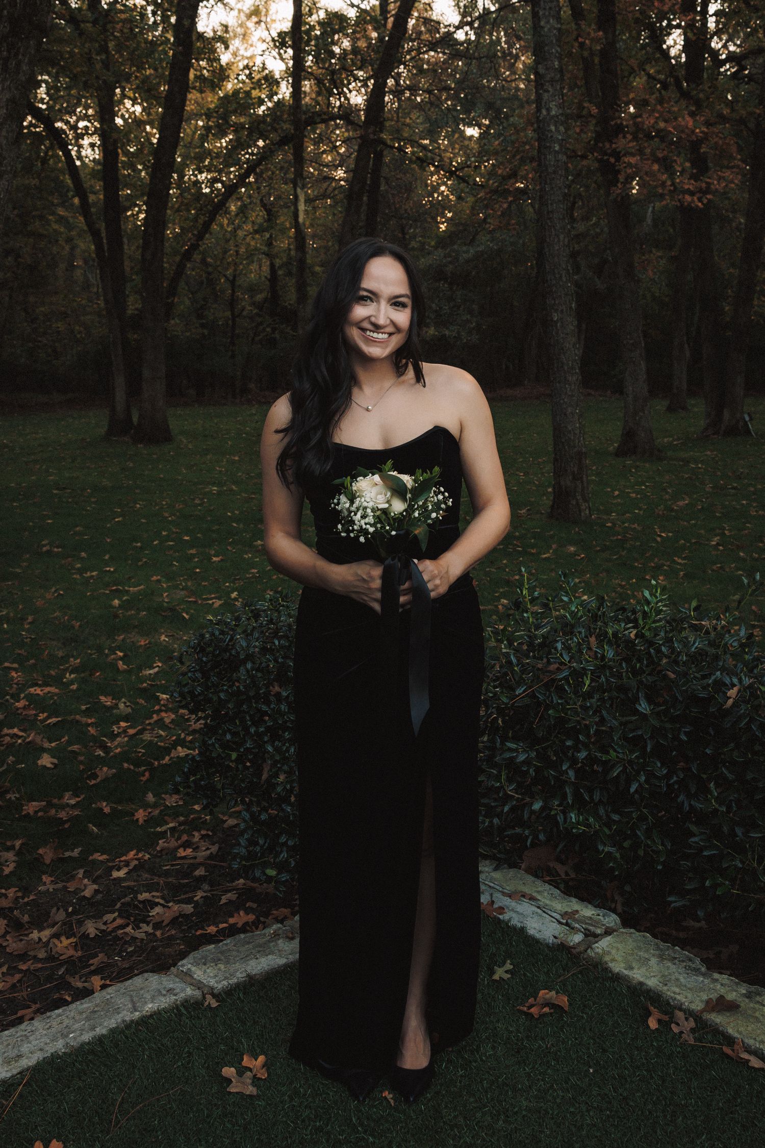 Bridesmaid in elegant black strapless gown holding white bouquet smiling in autumn garden.