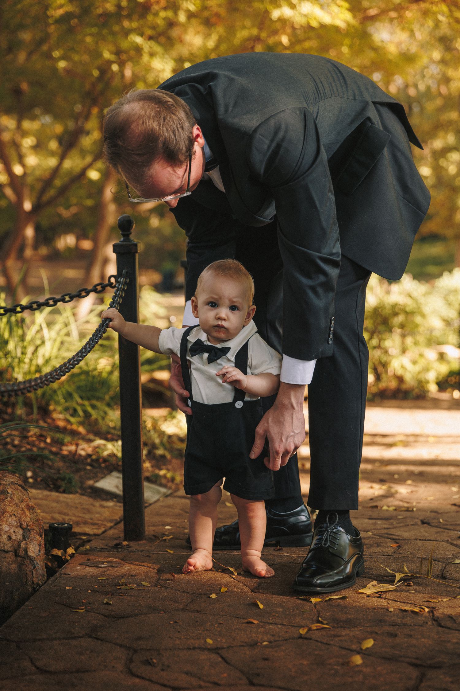 Man in dark suit helps a dressed-up baby boy stand on a stone garden path at a wedding.