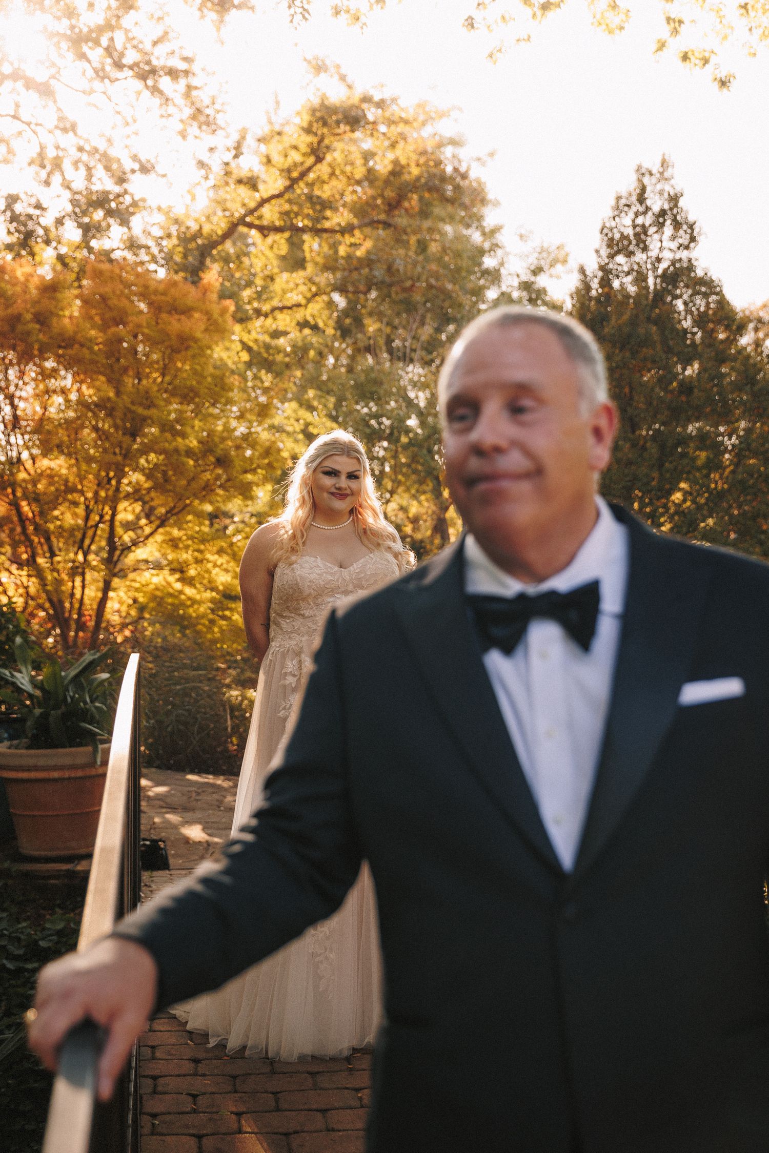 Bride in lace gown smiles at groom in black tuxedo during golden hour outdoor wedding ceremony.