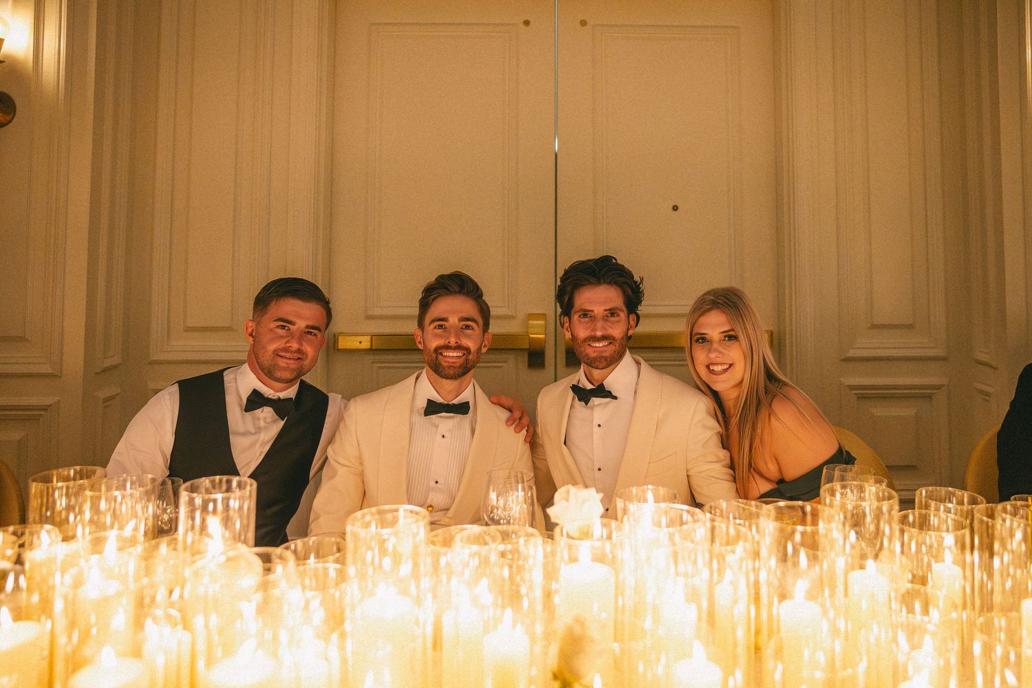 Two grooms in ivory tuxedos posing with friends surrounded by glowing candles at wedding reception.