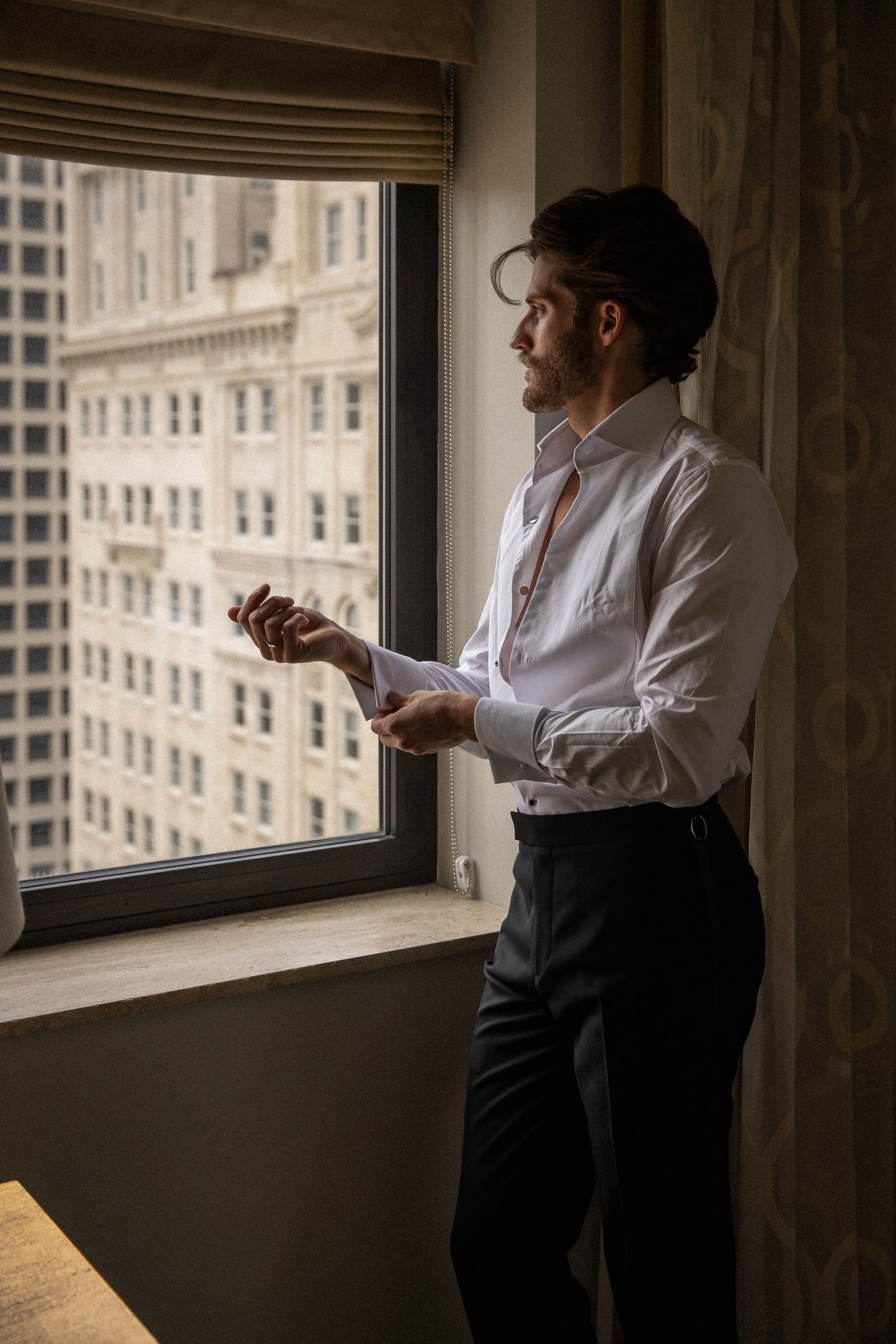 Handsome groom in white dress shirt buttoning cuffs by hotel window with city skyline view.