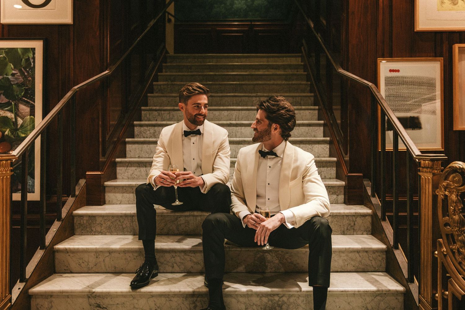 Two grooms in white tuxedos sitting on ornate staircase sharing a moment.