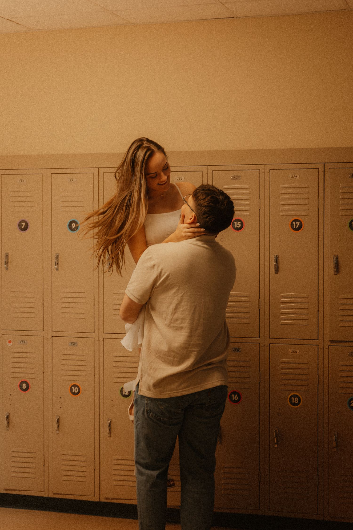A couple shares a romantic moment by school lockers, with the man lifting the woman in a warmly lit hallway.