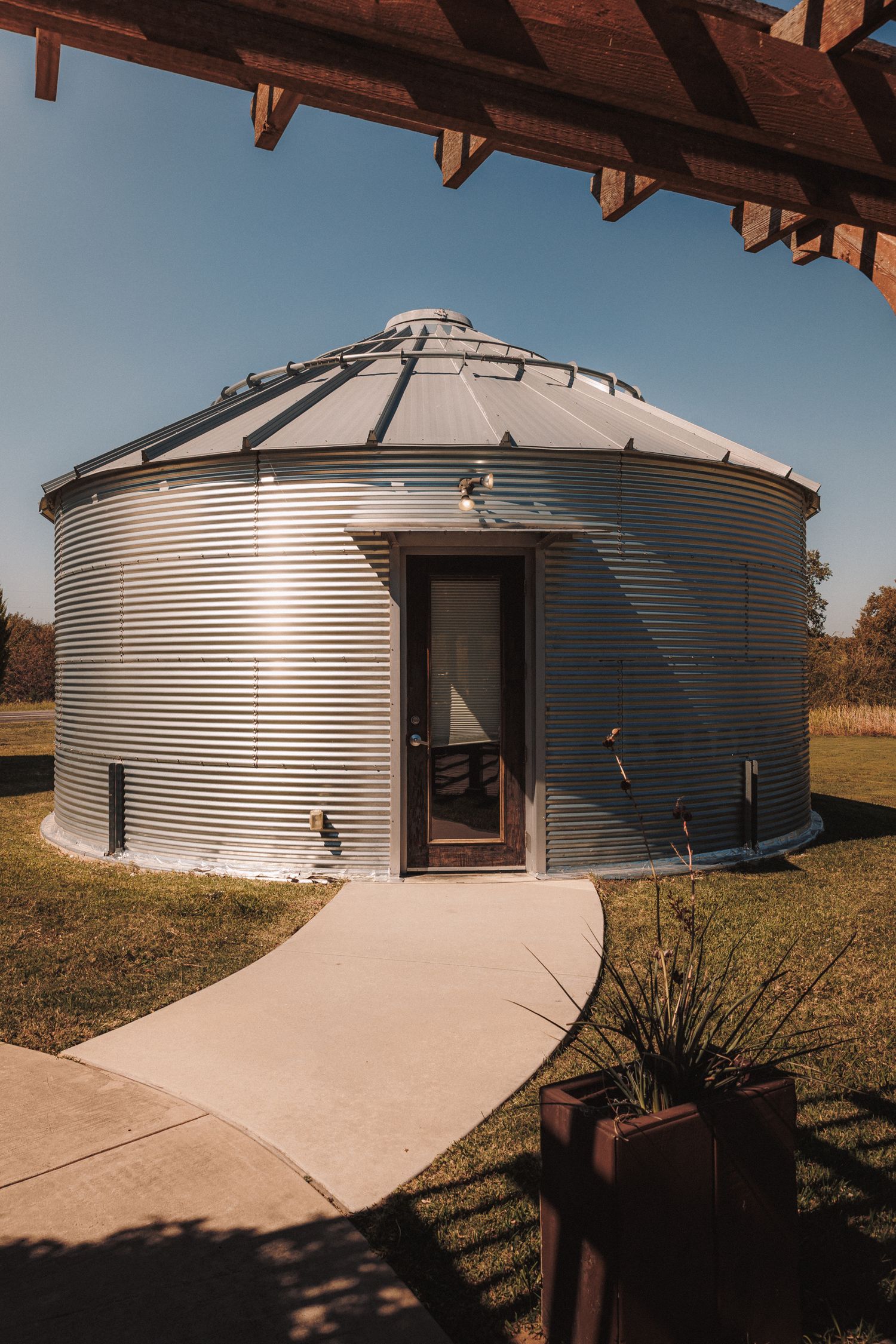 A converted grain silo with corrugated metal walls and a wooden door serves as a unique rustic dwelling on a sunny day.