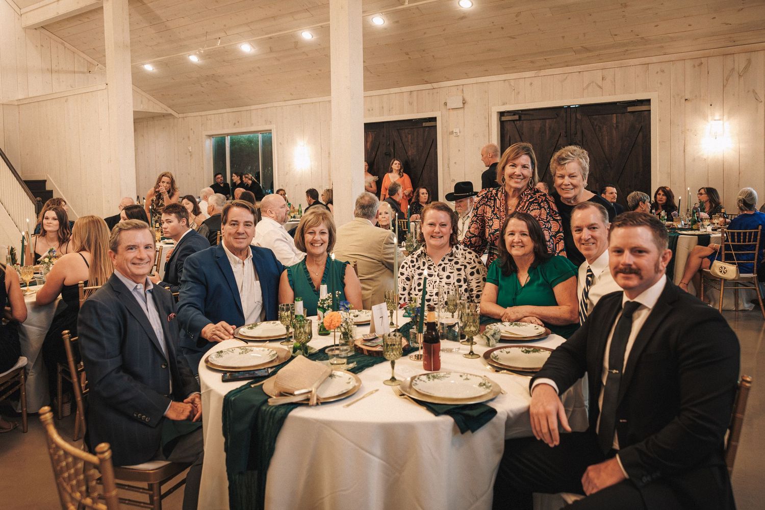 Wedding guests smile and pose together at a round reception table decorated with green runners and floral centerpieces.