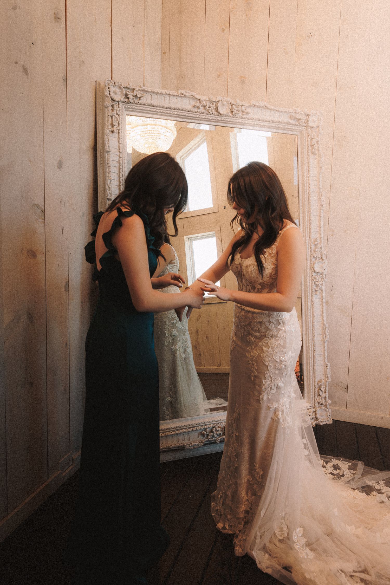 Bridesmaid in teal dress helping bride in lace gown prepare in front of ornate white mirror.