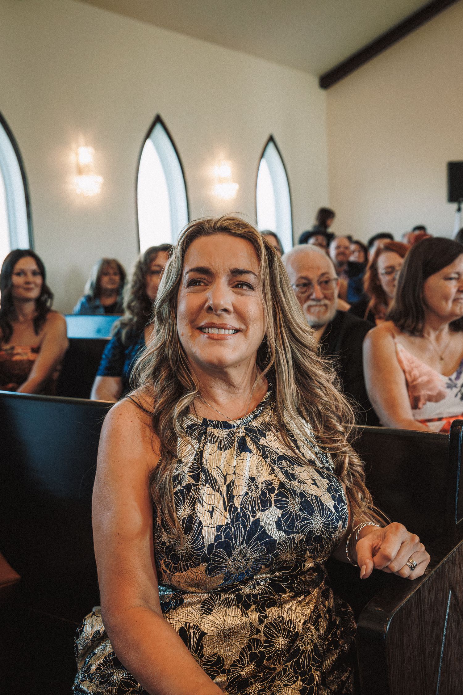 Smiling woman with blonde highlights seated in a church pew surrounded by wedding guests.