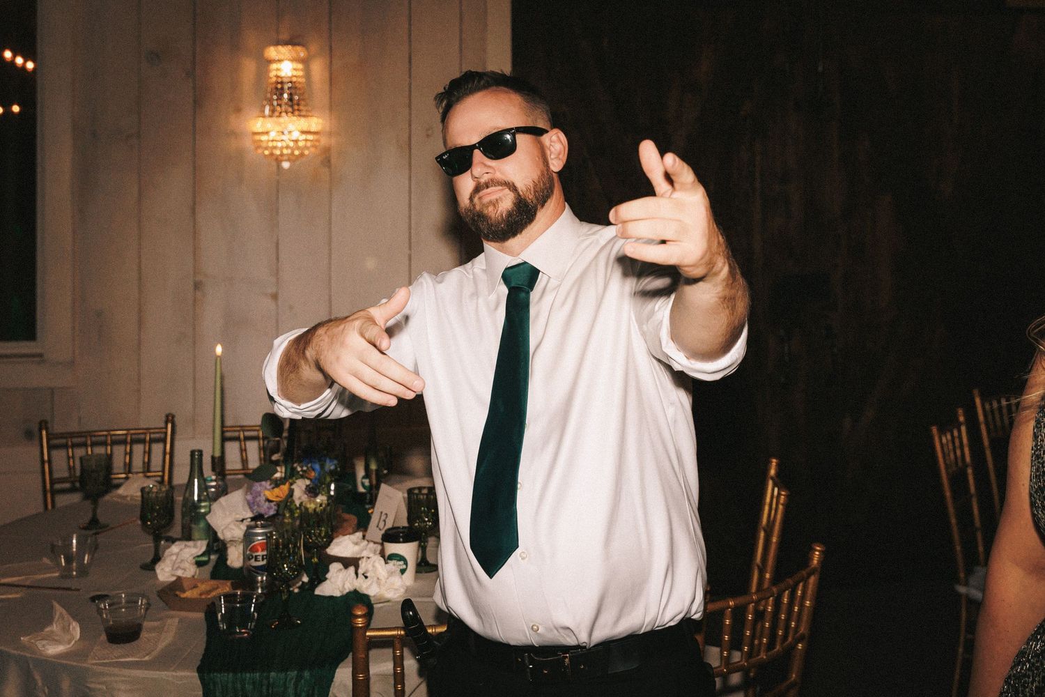 Energetic wedding guest in sunglasses and green tie dancing at a candlelit reception table.