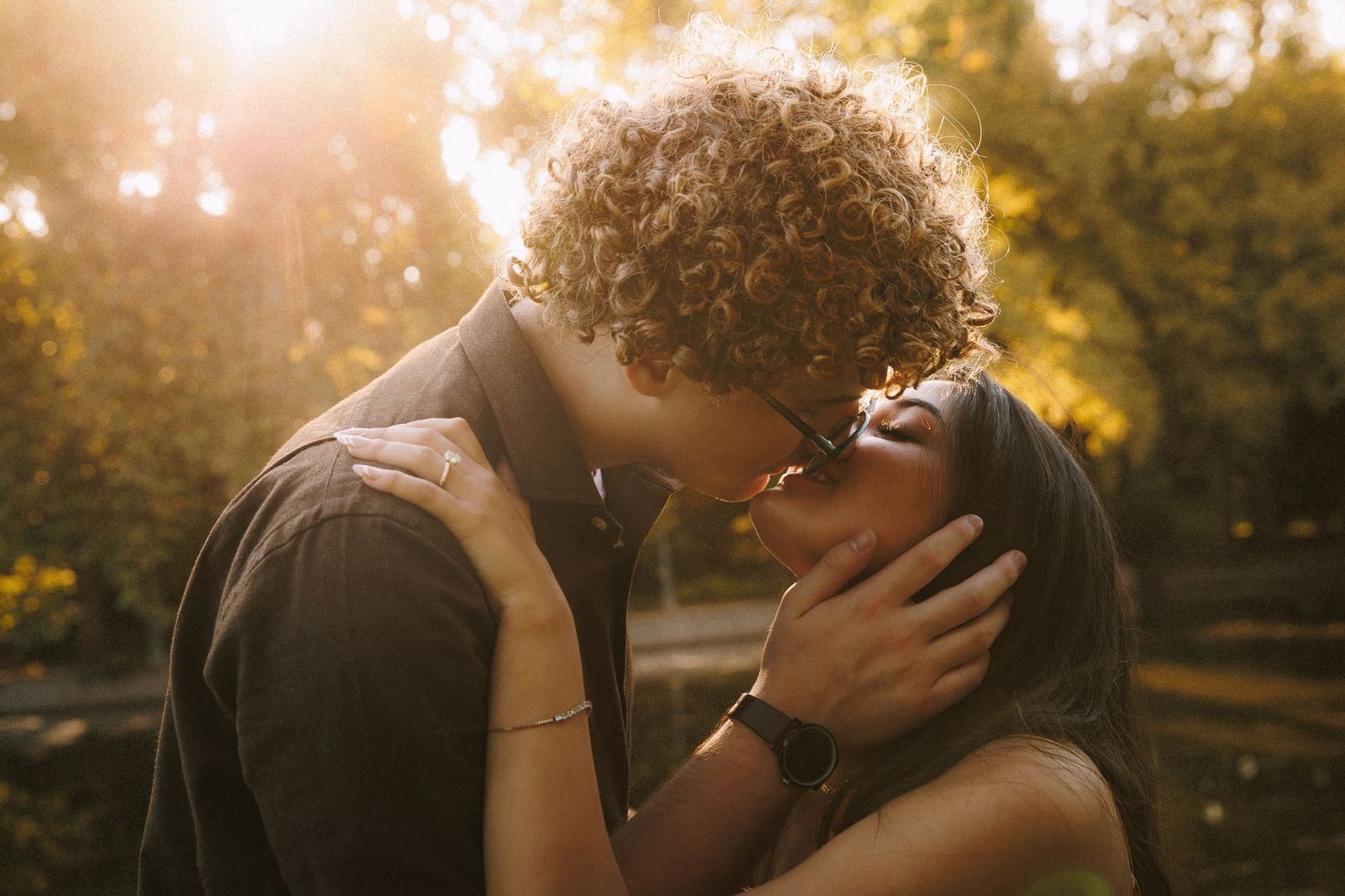 A romantic couple embraces during a golden sunset in a park setting with trees in the background.