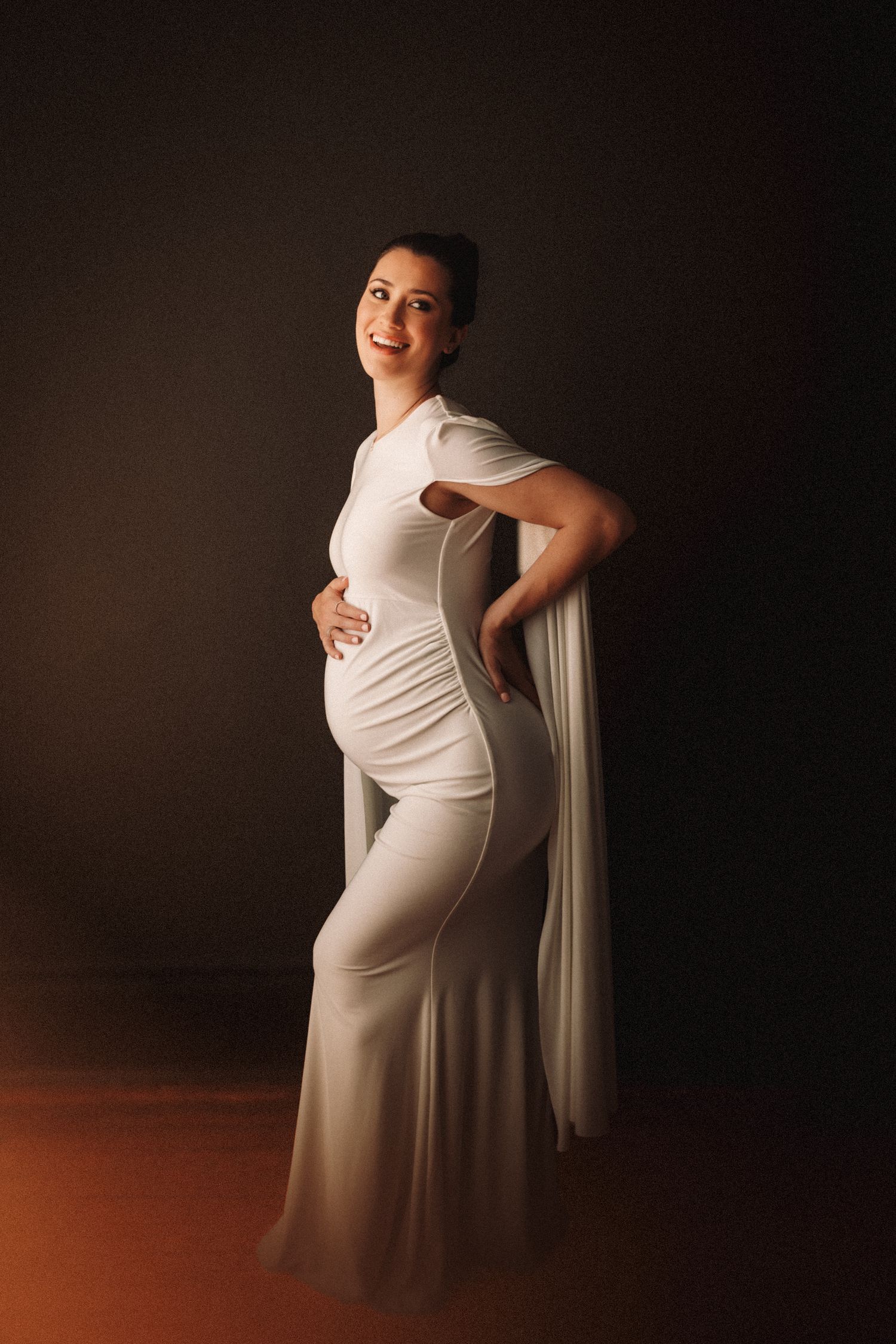 Elegant maternity photos with a figure in a white dress posing against a dark background with warm lighting.