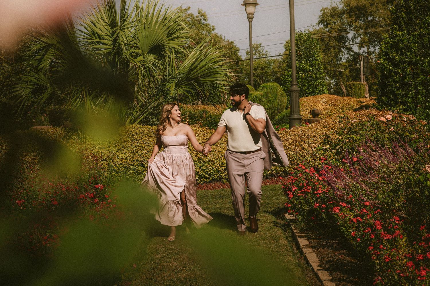 A couple walks through a lush garden path with blooming red roses and greenery during a sunny afternoon.