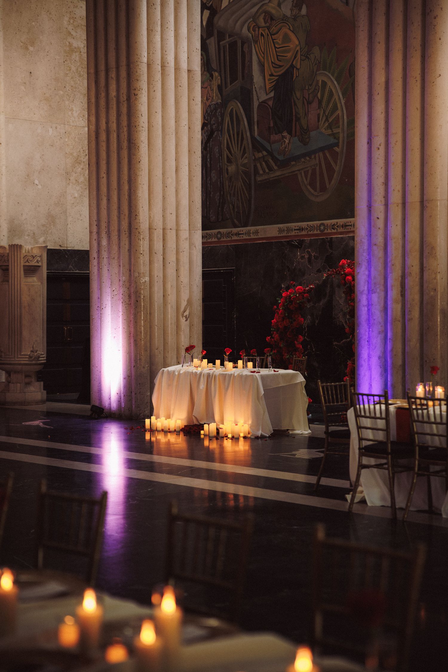 Elegant candlelit wedding reception in grand hall with white draped sweetheart table, red floral arrangements, and tall marble columns.