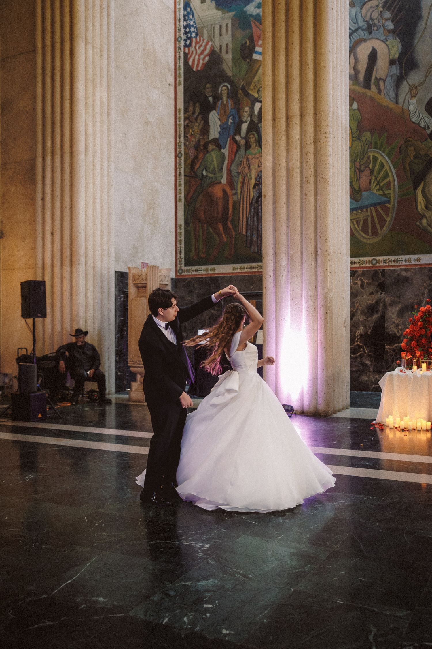 Bride and groom sharing first dance in grand hall with large murals and marble floors.