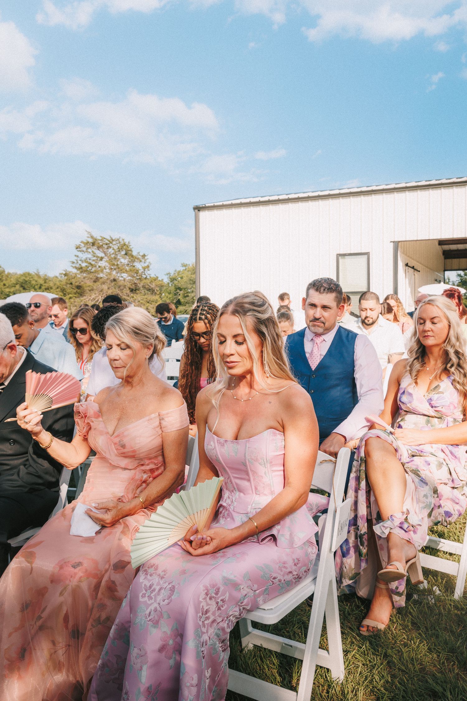 Wedding guests in pink dresses seated on white chairs at an outdoor ceremony.