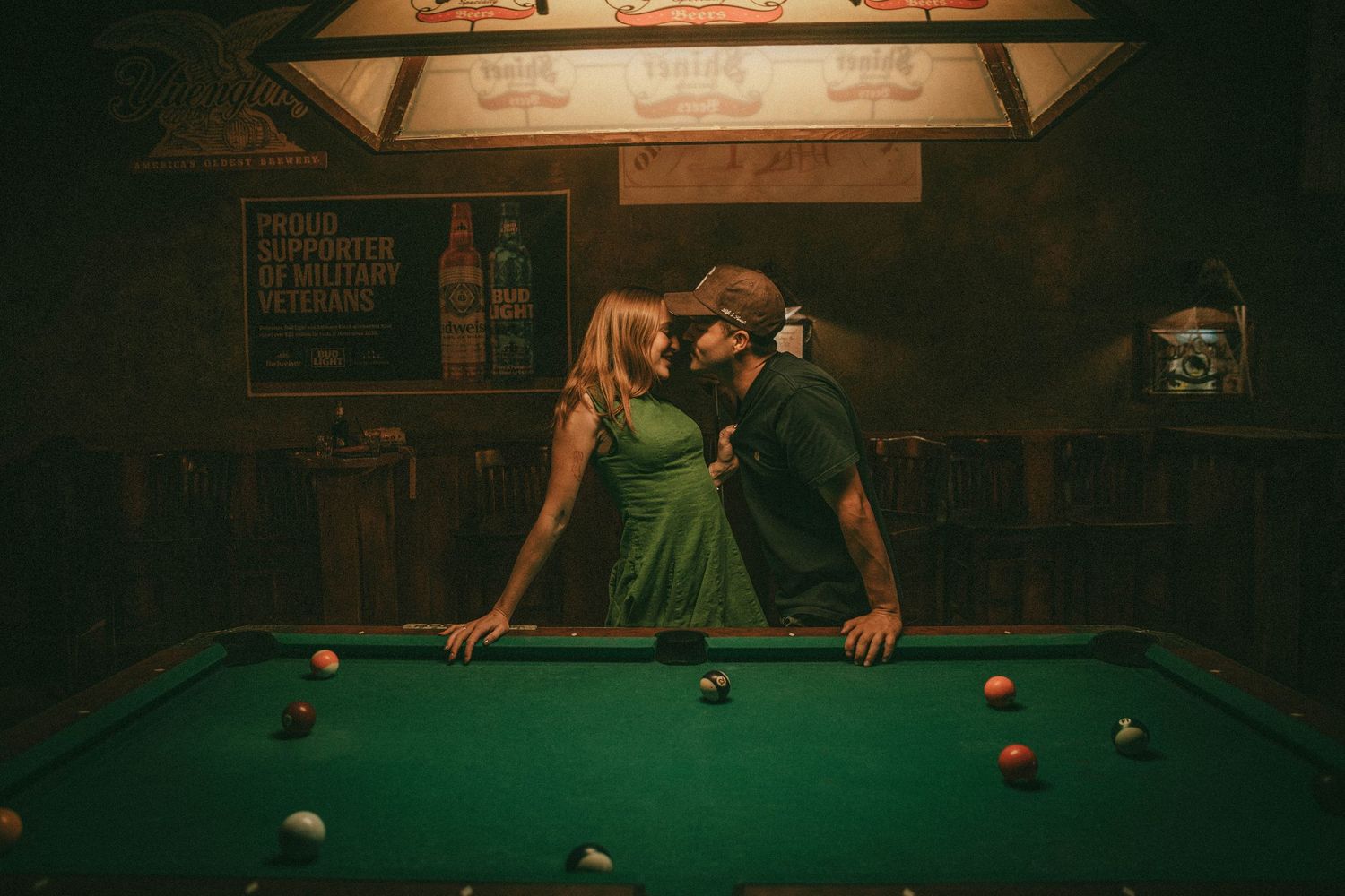 Players lean over a pool table in a dimly lit bar setting during a game of billiards.