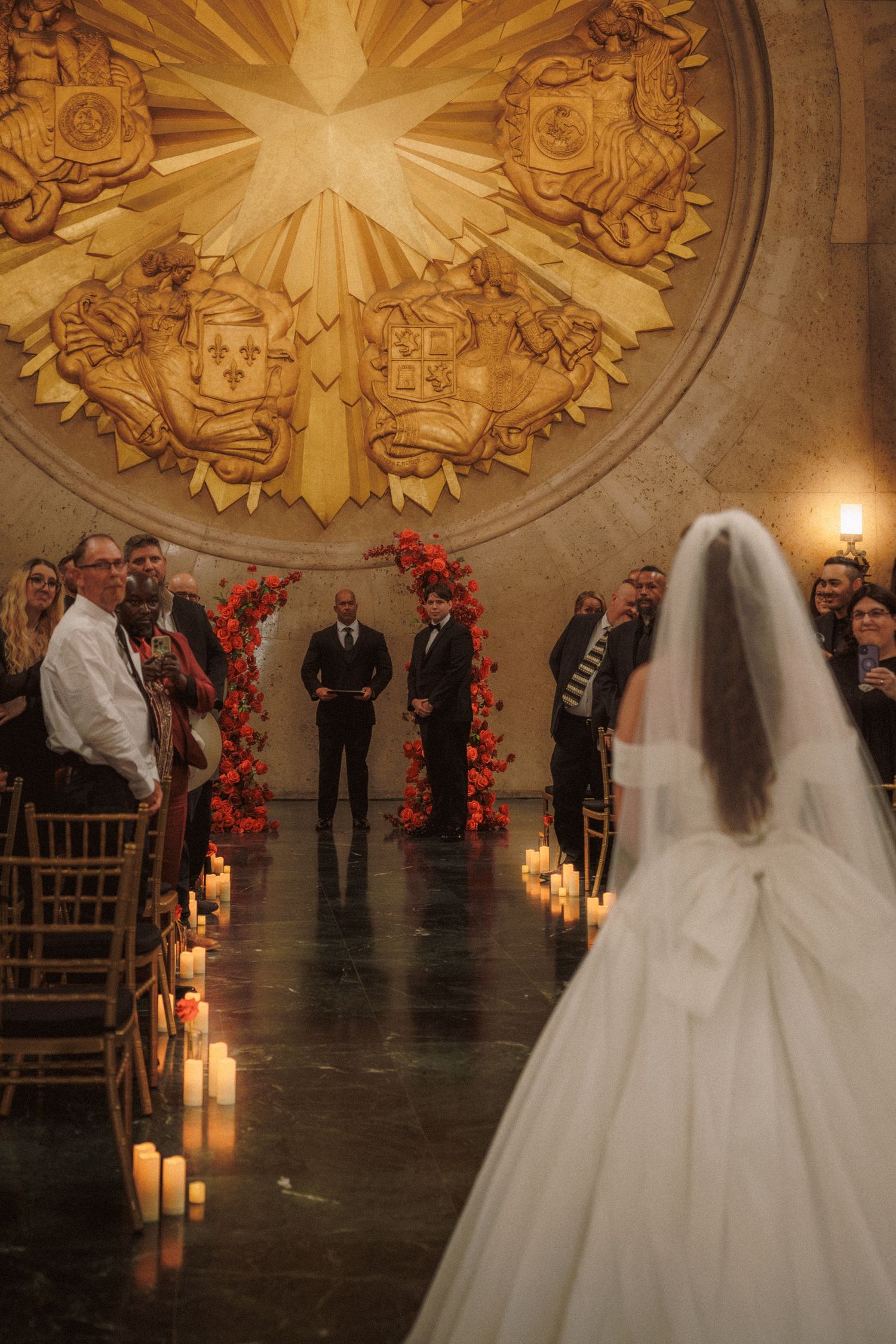 Bride walks down candlelit aisle toward floral arch in ornate golden domed wedding venue.