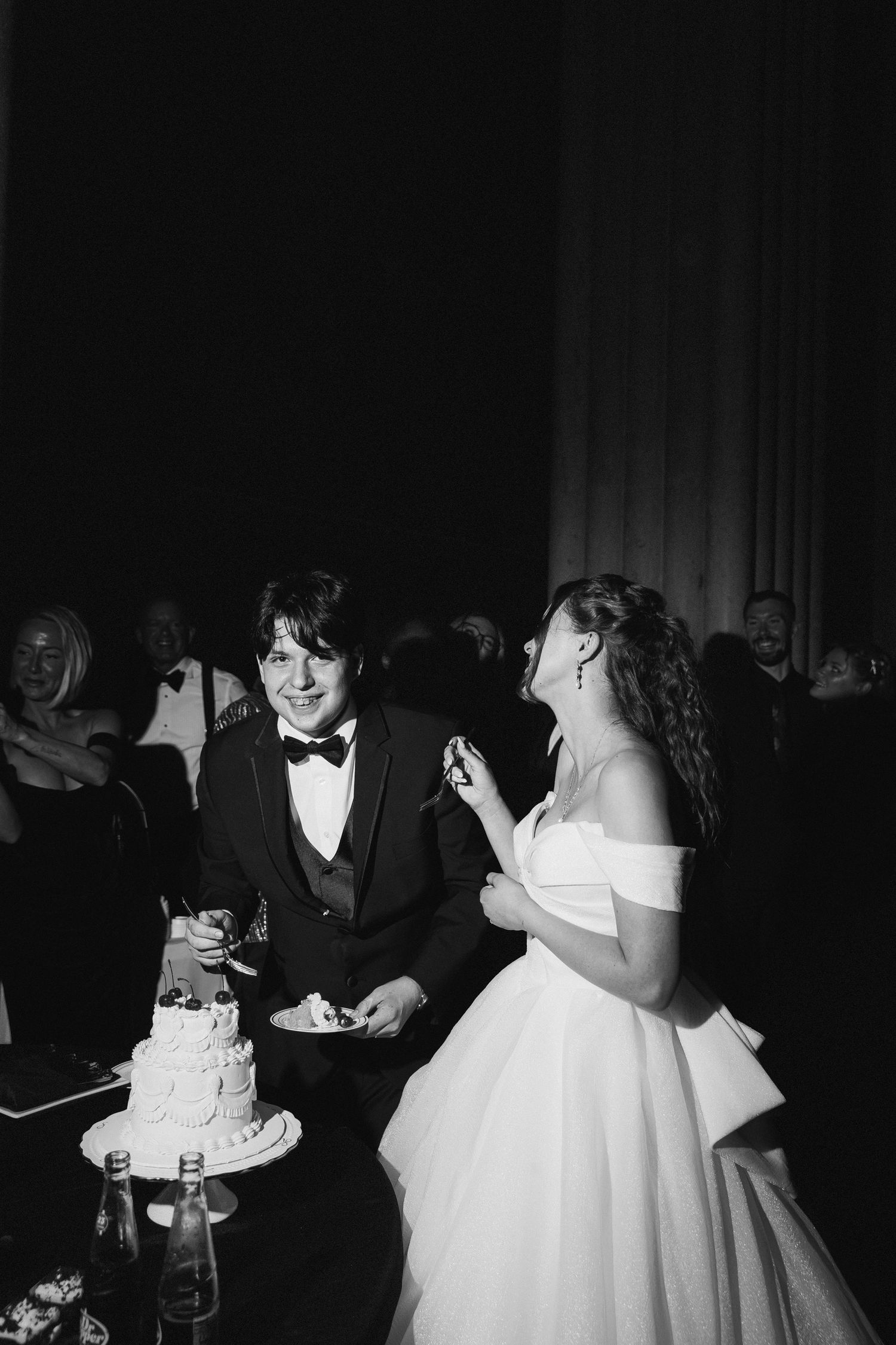 Black and white photo of bride and groom laughing while cutting wedding cake at reception.