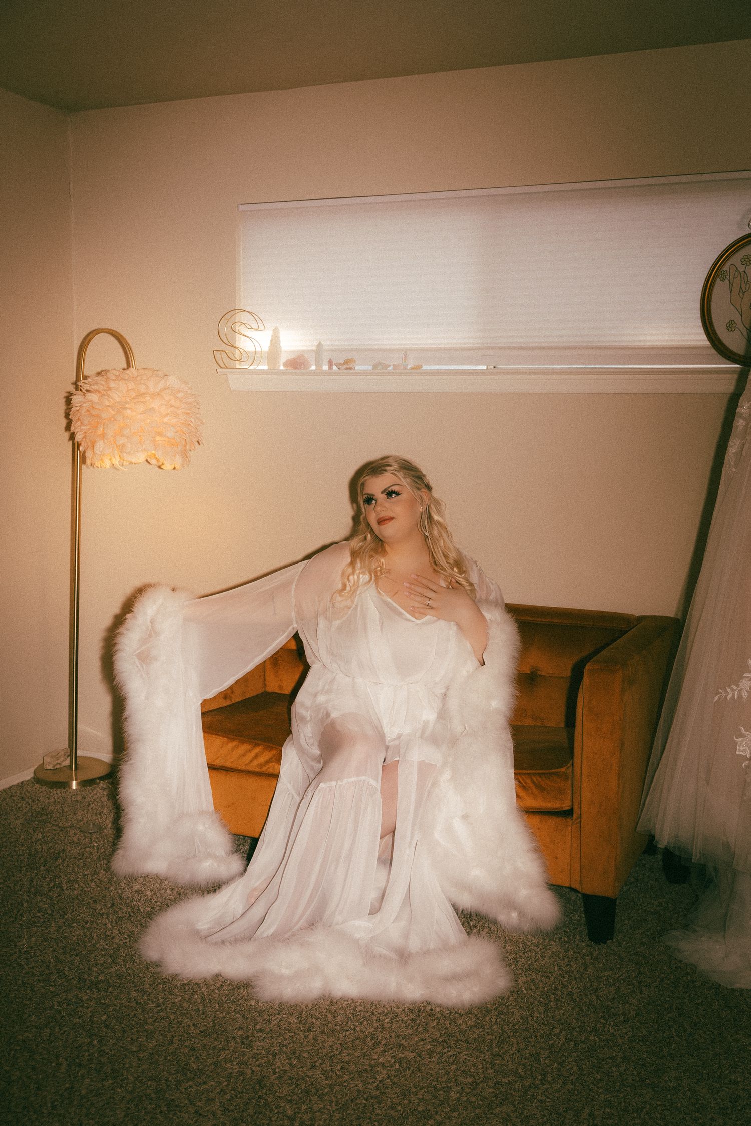 Bride posing in a flowing white feather-trimmed bridal robe beside a gold velvet sofa.