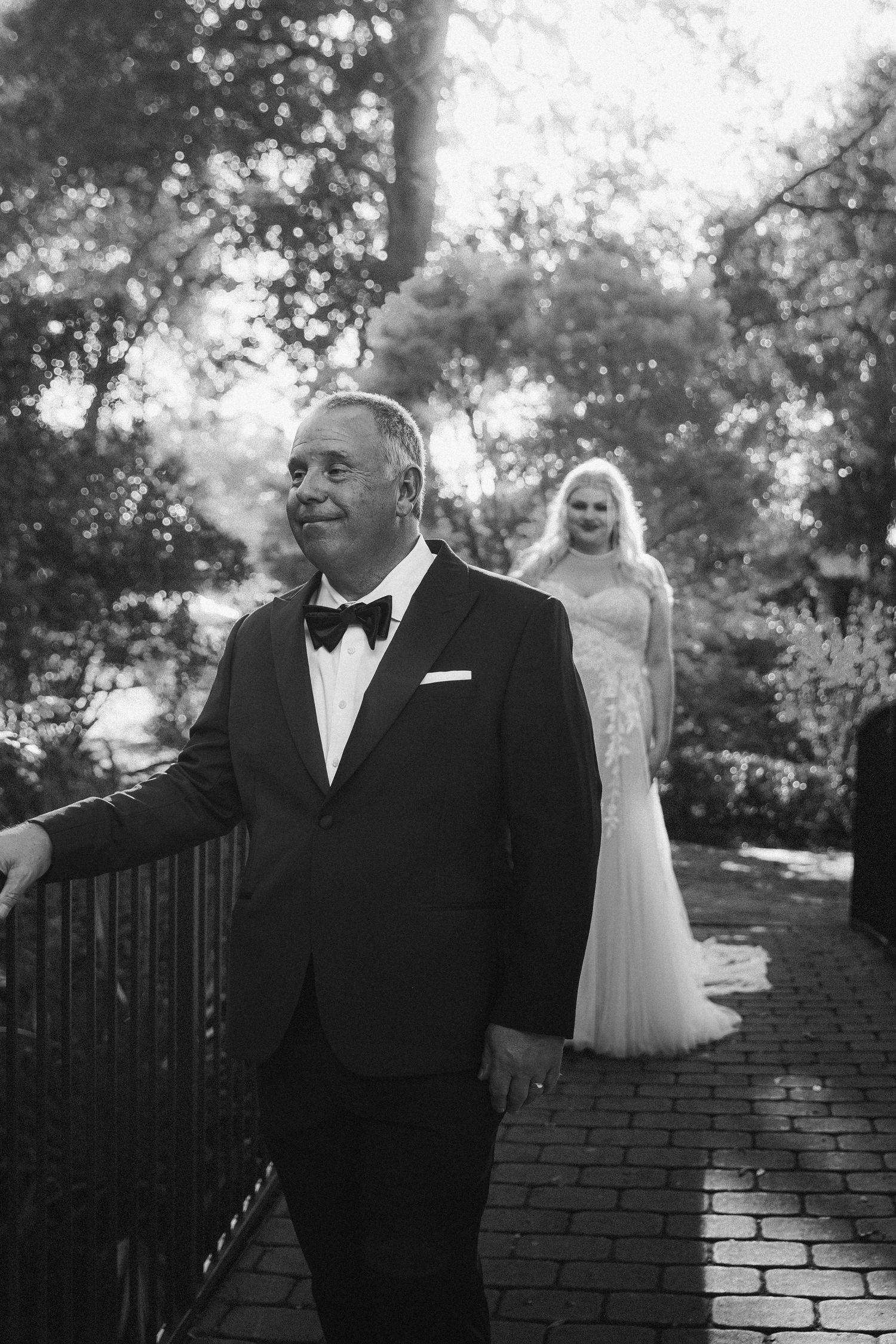 Black and white photo of groom in tuxedo with bride in wedding gown approaching on brick path.