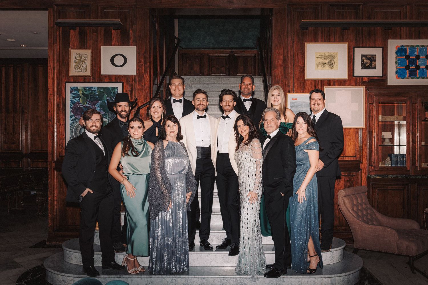 Elegant wedding party group photo with guests in formal attire posing together in a warmly lit venue.